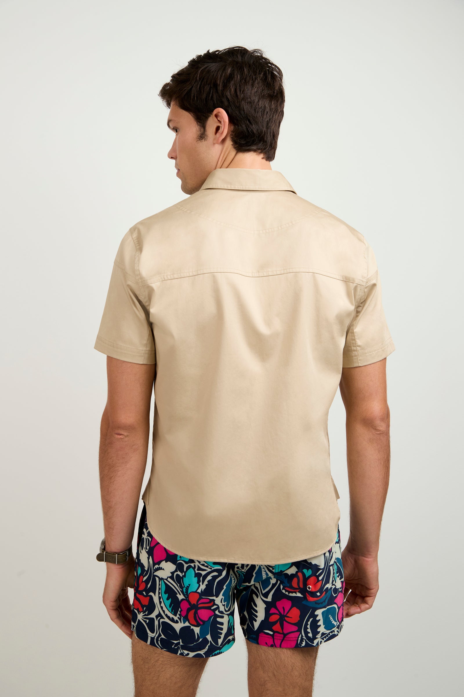 A man stands with his back to the camera, wearing the Shelton Short-Sleeve Shirt and colorful floral shorts against a plain light background.