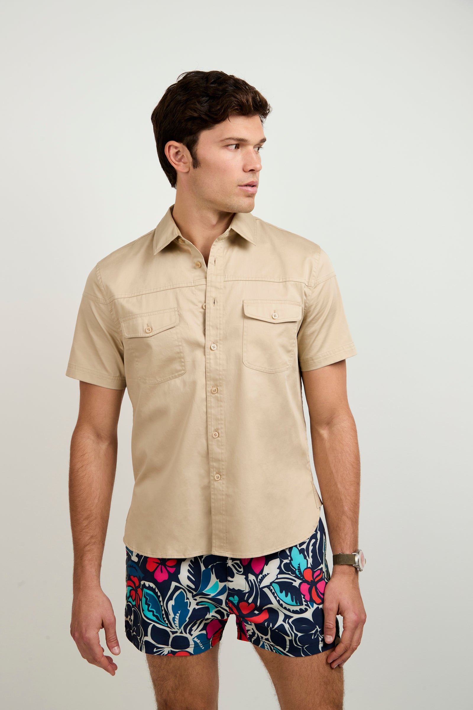 A man wears the Shelton Short-Sleeve Shirt in beige and colorful floral shorts, standing against a plain light background while looking to his left with a neutral expression.