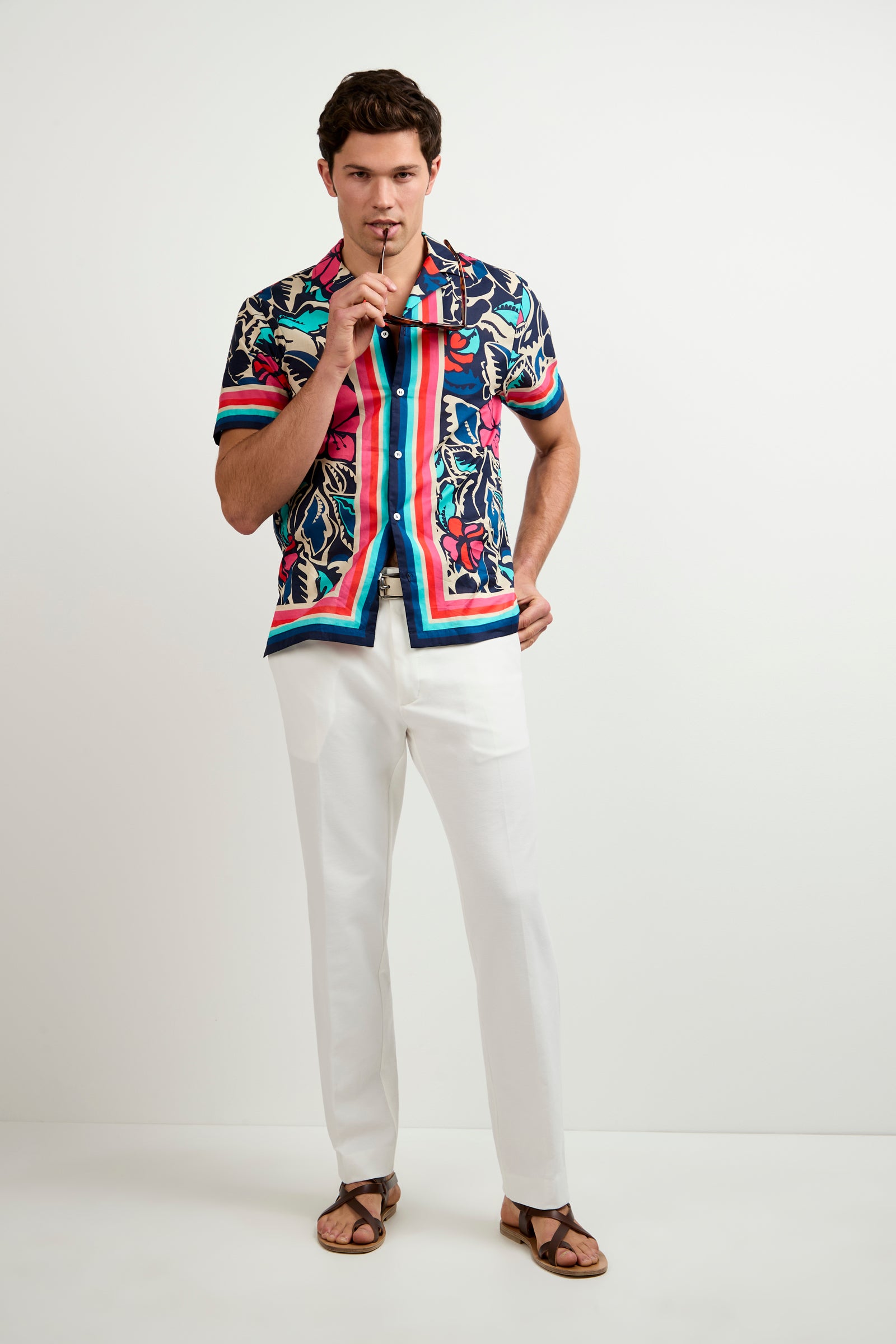 A man stands against a plain background wearing the Josue Short-Sleeve Shirt - Copacabana, featuring bold colorful patterns. He pairs it with white pants, brown sandals, and holds sunglasses near his mouth while looking forward.