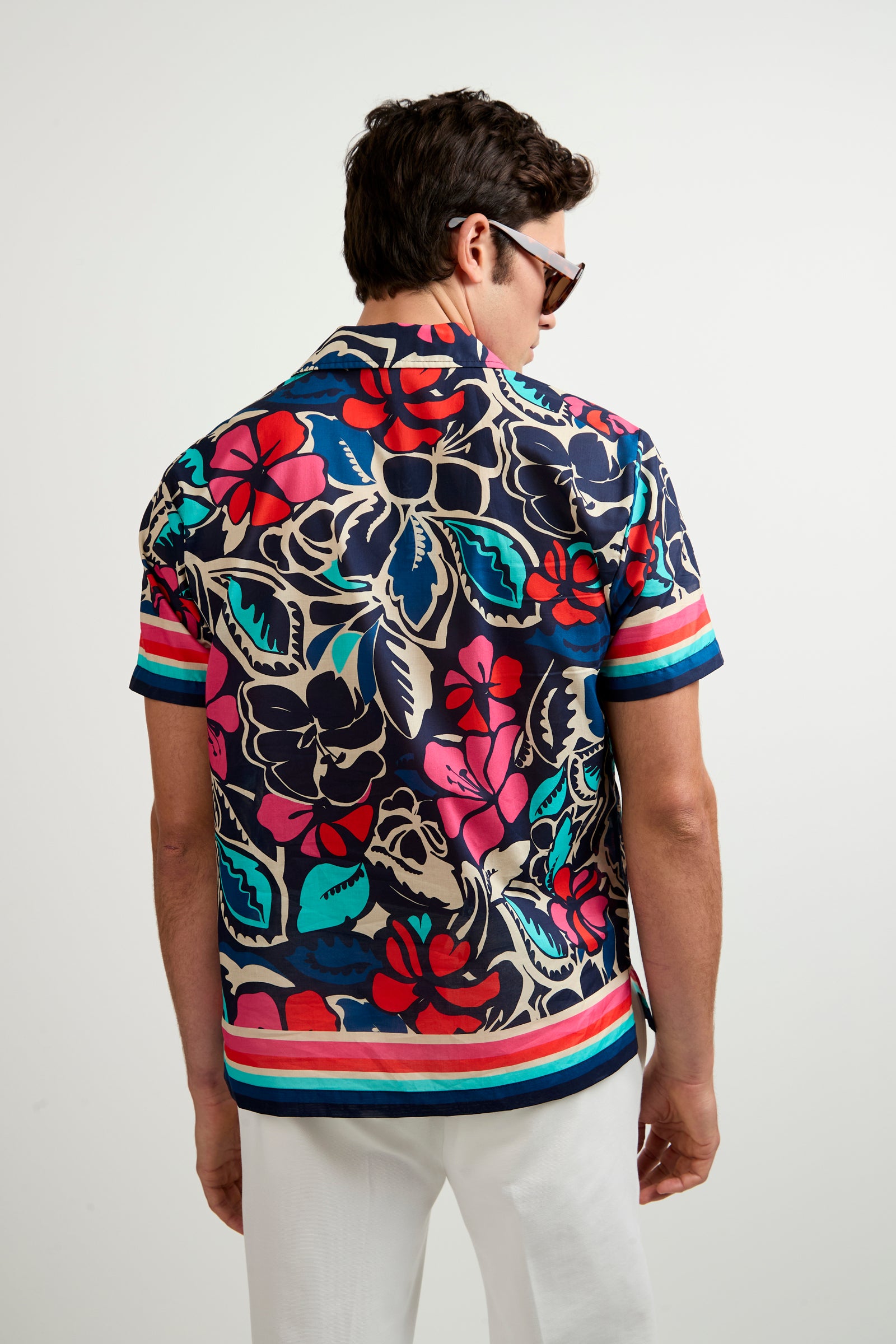 A man wearing the Josue Short-Sleeve Shirt - Copacabana, featuring a vibrant red, blue, and cream floral pattern, and white pants stands facing away. He also has sunglasses on.