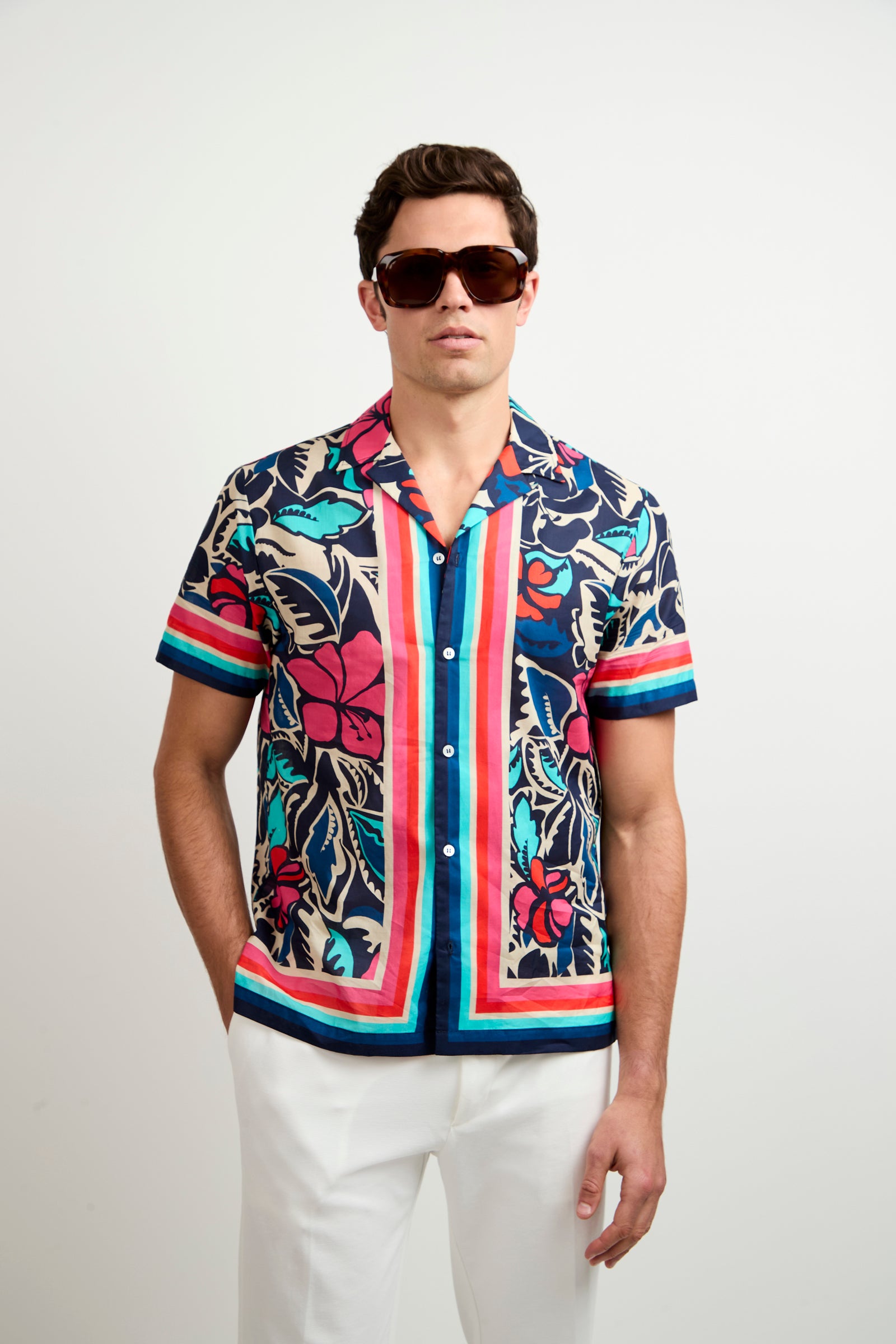A man stands against a plain white background wearing large dark sunglasses, white pants, and the Josue Short-Sleeve Shirt - Copacabana, a colorful button-up shirt with a bold floral and stripe pattern.