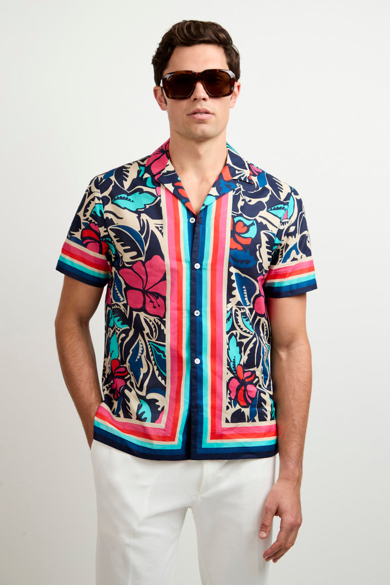 A man wearing the Josue Short-Sleeve Shirt - Copacabana, featuring a vibrant floral and stripe pattern, paired with dark sunglasses and white pants, stands against a plain light background.