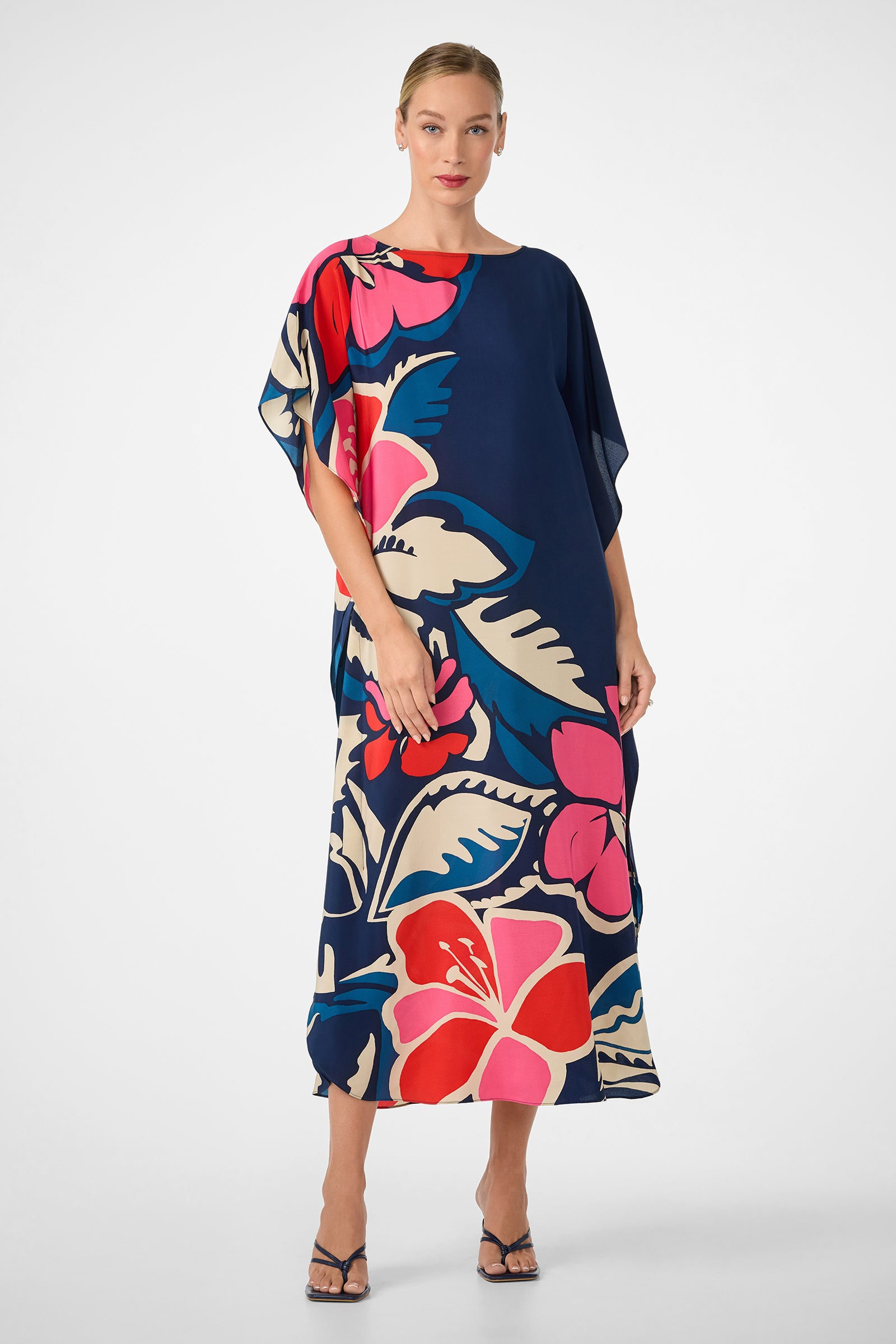 A woman wears the Jalani Maxi Caftan Dress, an ankle-length navy blue dress with oversized pink, red, and cream tropical florals, paired with strappy heels against a plain white background.