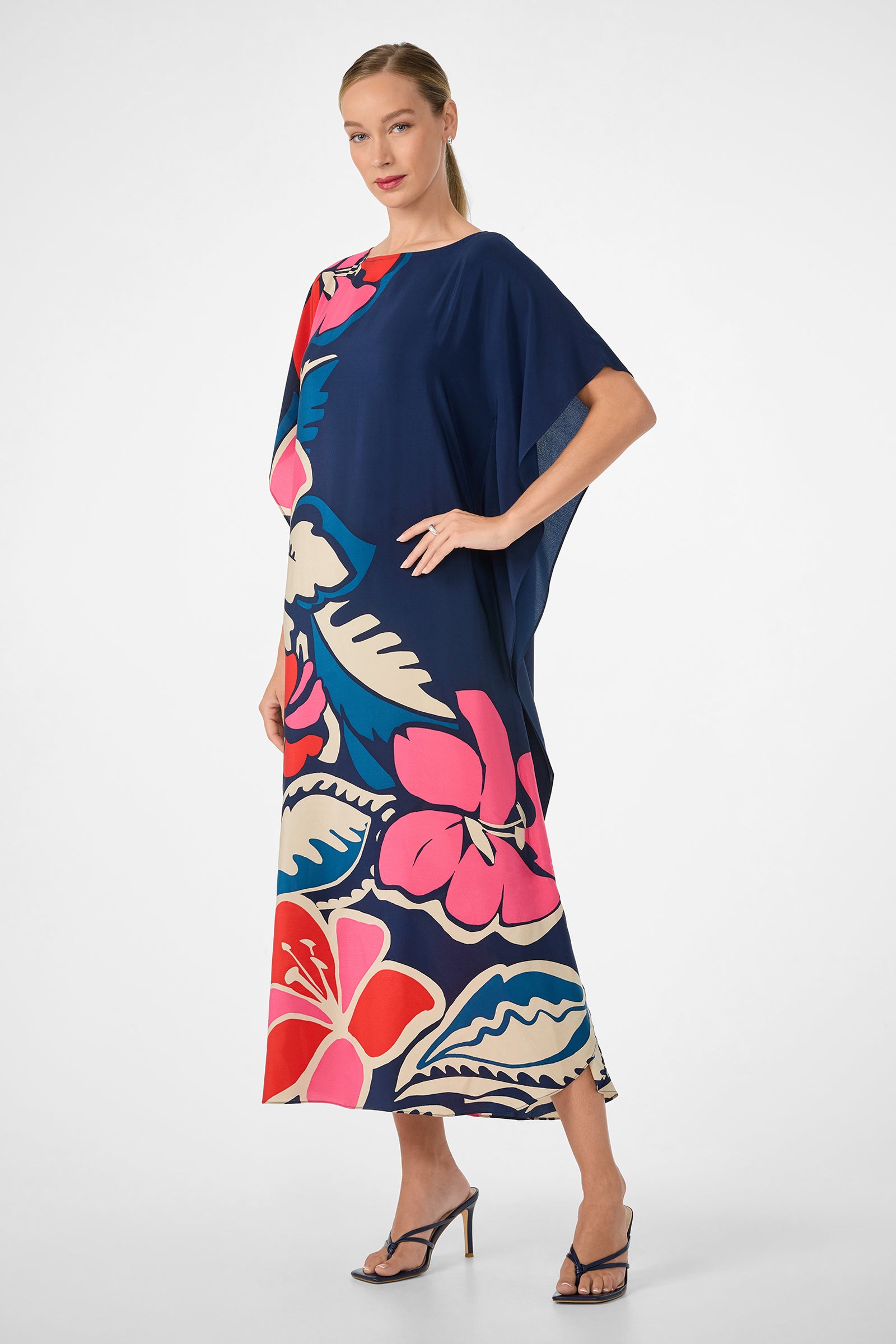 A woman models the Jalani Maxi Caftan Dress, a loose-fitting navy blue maxi with bold pink, red, white, and blue floral prints. She pairs it with black heeled sandals and a sleek pulled-back hairstyle.