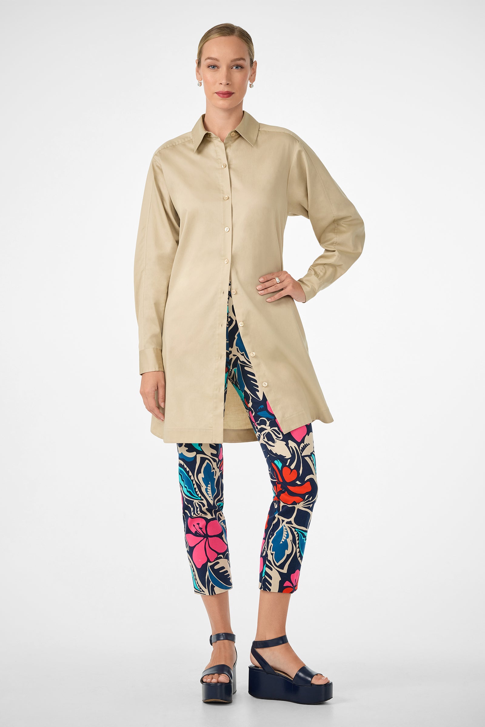 A woman stands facing forward in the Marx Dress, a long beige button-up shirt. She pairs it with colorful floral leggings, has her hair tied back, and wears navy platform sandals against a plain white background.