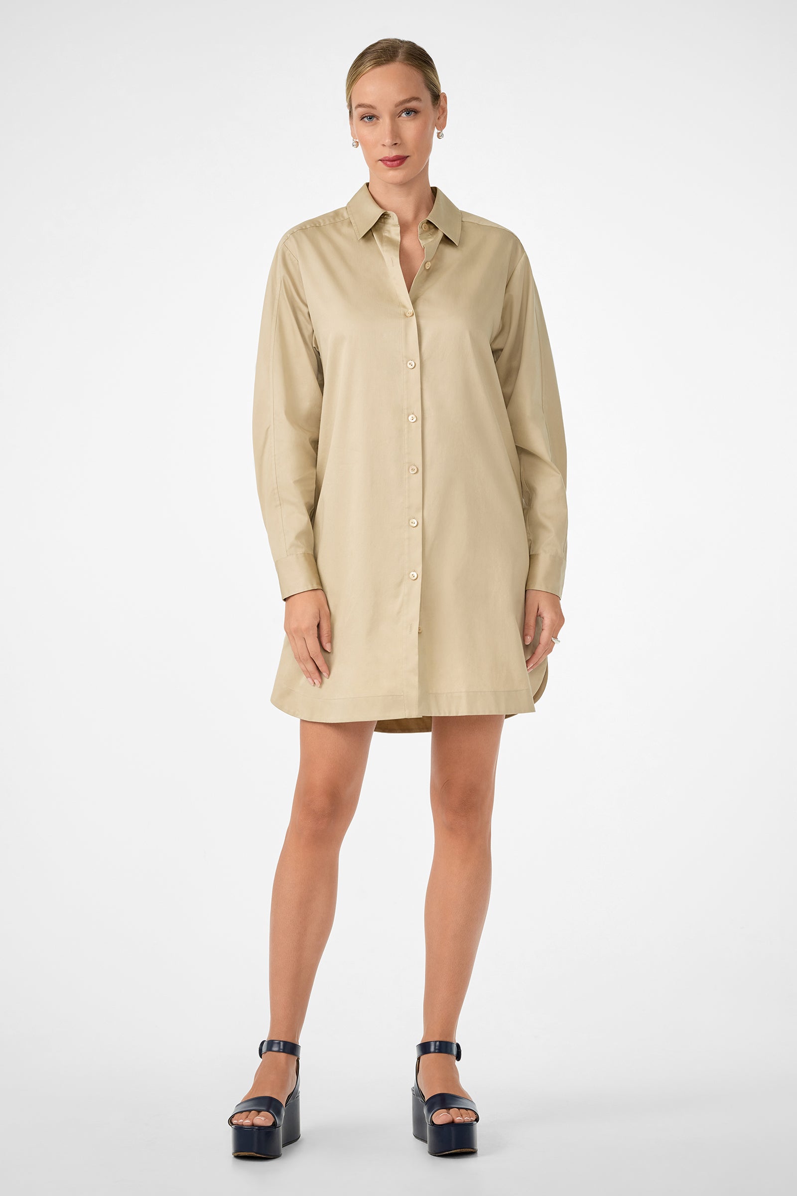A woman with light skin and tied-back hair stands facing forward, wearing the Marx Dress—a light beige, long-sleeve, collared shirt dress—paired with black platform sandals, against a plain white background.
