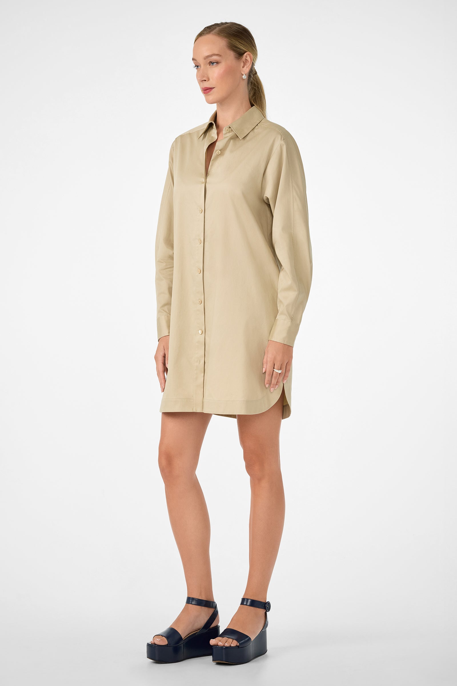 A woman stands against a white background wearing the beige Marx Dress, a long-sleeve, button-up shirt dress, paired with black platform sandals. Her hair is pulled back and she is facing slightly to the left.
