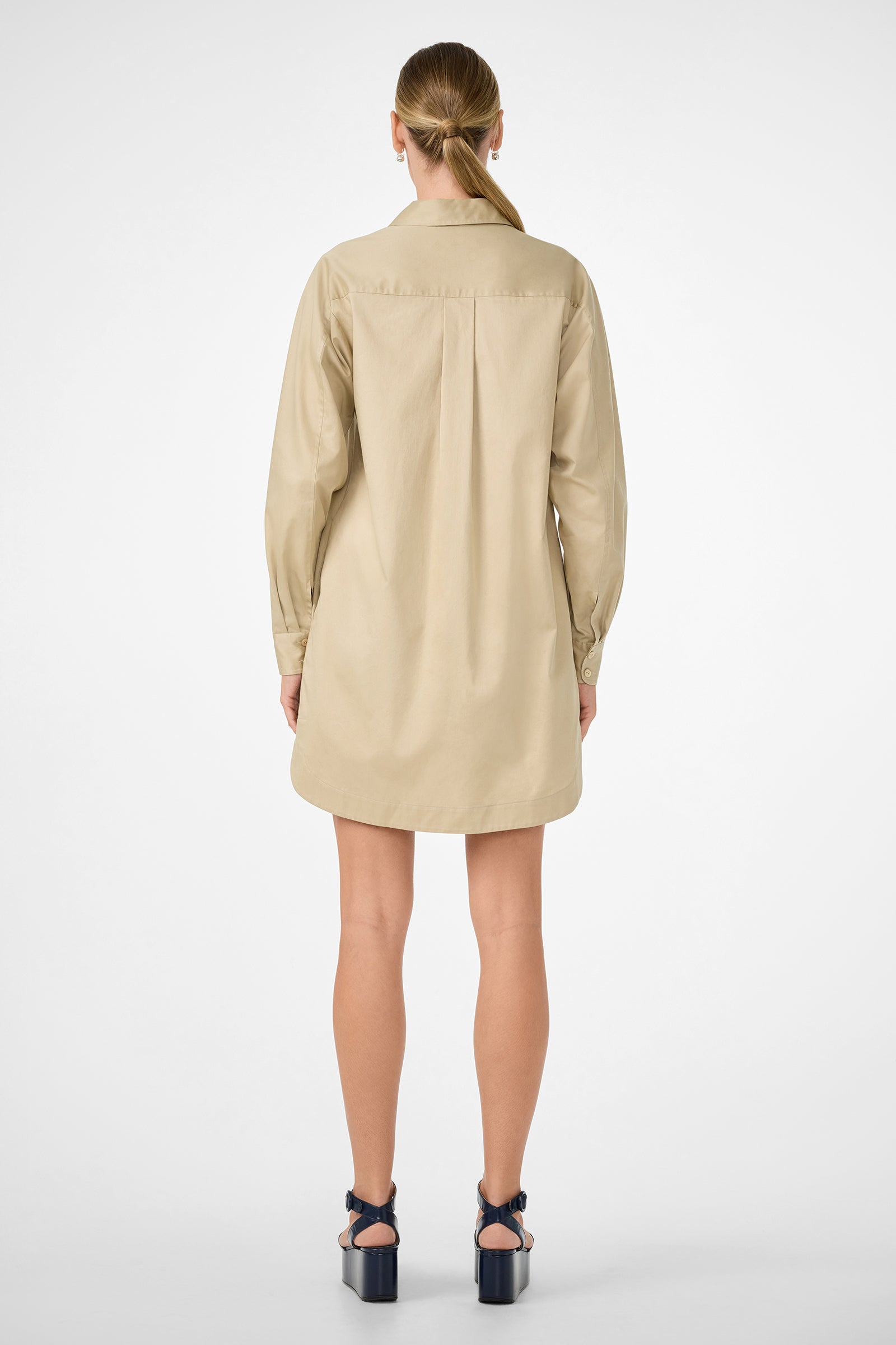 A woman stands facing away in the Marx Dress, a long-sleeved light beige shirt dress, with her light brown hair tied back in a low ponytail and wearing dark platform heels against a plain white background.