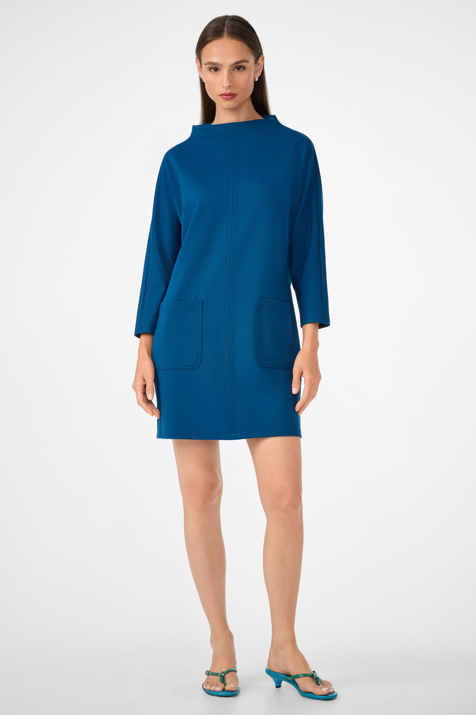 A woman with straight brown hair and a neutral expression stands against a white background, wearing the Caracas Dress—a teal, long-sleeve, knee-length dress with front pockets—and green open-toe heeled sandals.