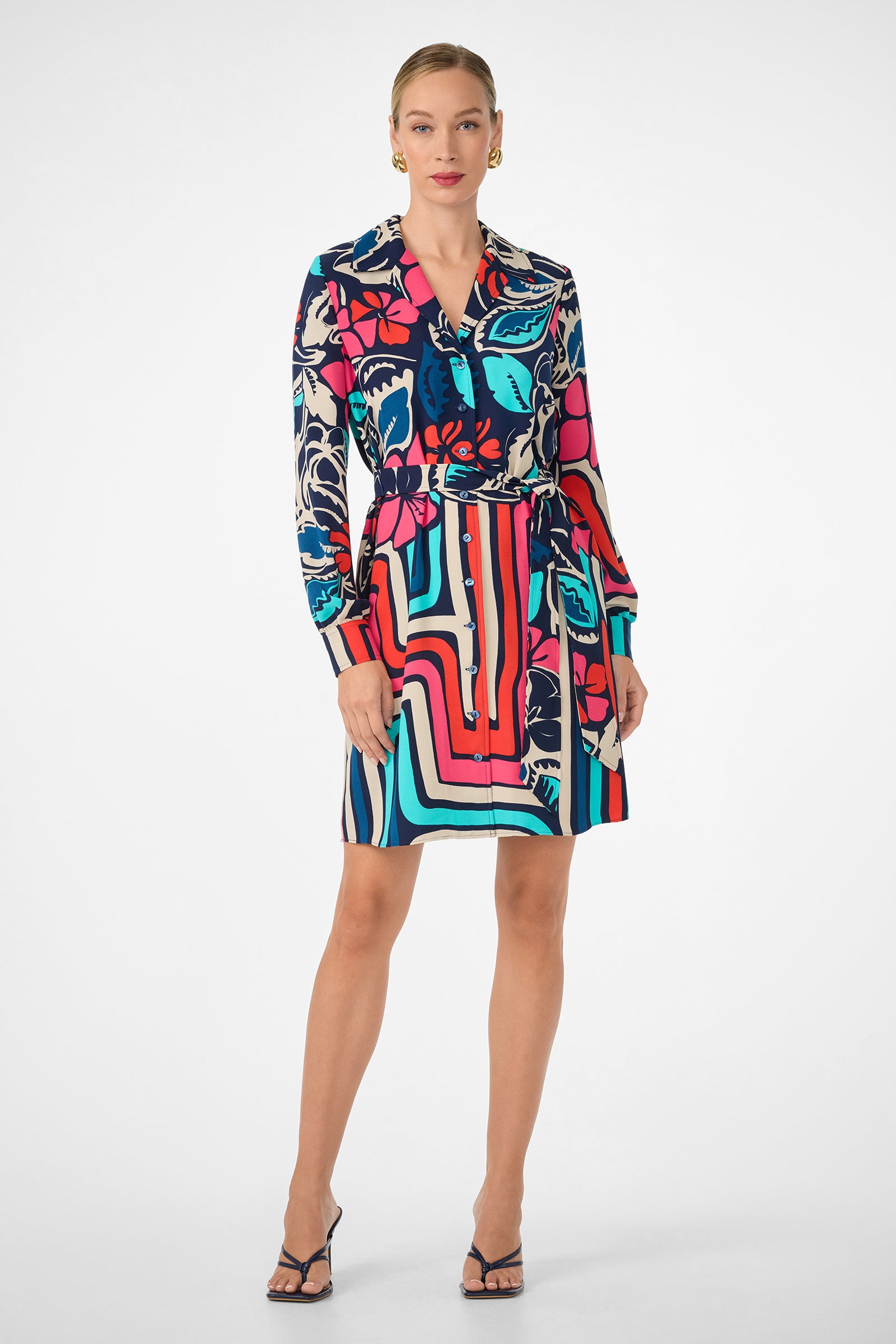 A woman faces forward, wearing the Flamenco Dress—a vibrant, long-sleeve button-up with bold red, blue, black, and beige abstract patterns. Her hair is tied back and she pairs the look with strappy high-heeled sandals.