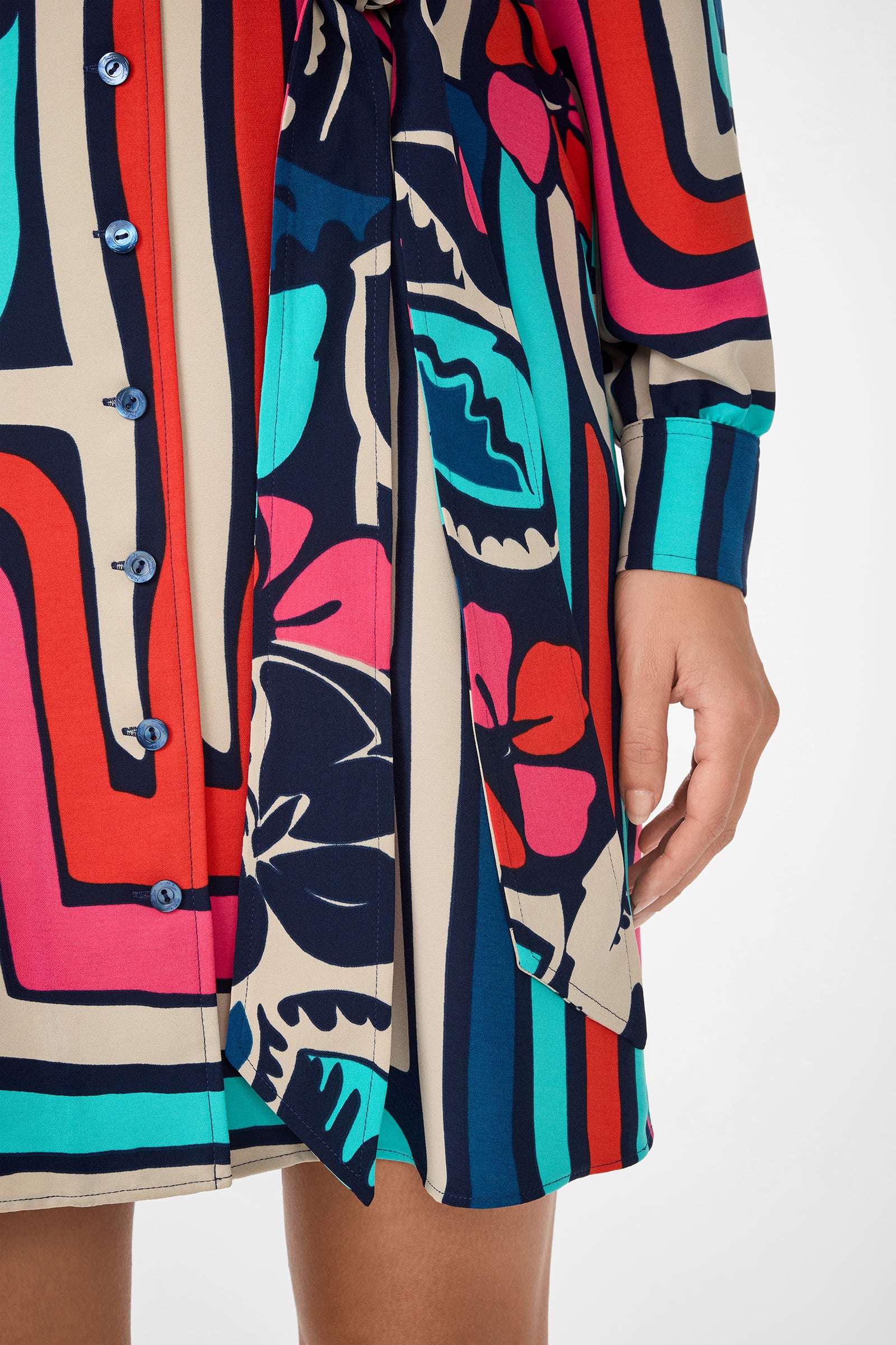 A person wearing the Flamenco Dress, a buttoned dress with bold, colorful abstract patterns in red, blue, teal, and beige, long sleeves, and a fabric waist tie. The photo highlights the lower torso and hand.