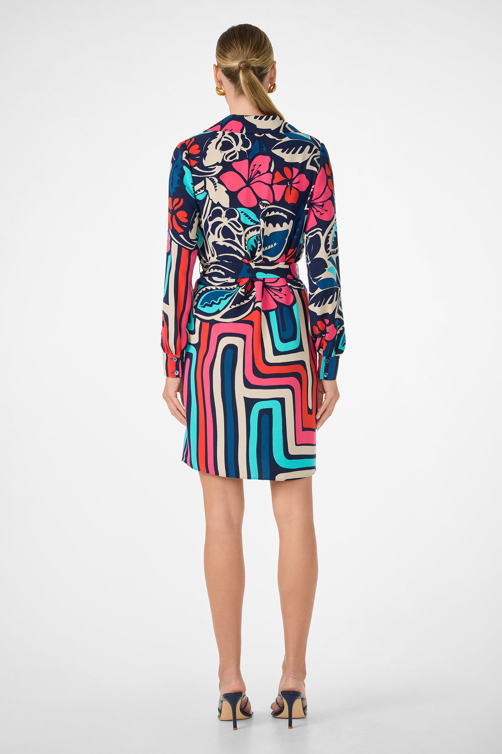 A woman stands facing away, wearing the Flamenco Dress—an above-the-knee, long-sleeve piece with bold, colorful floral and geometric patterns. She pairs it with open-toe heels and styles her hair in a neat low bun.