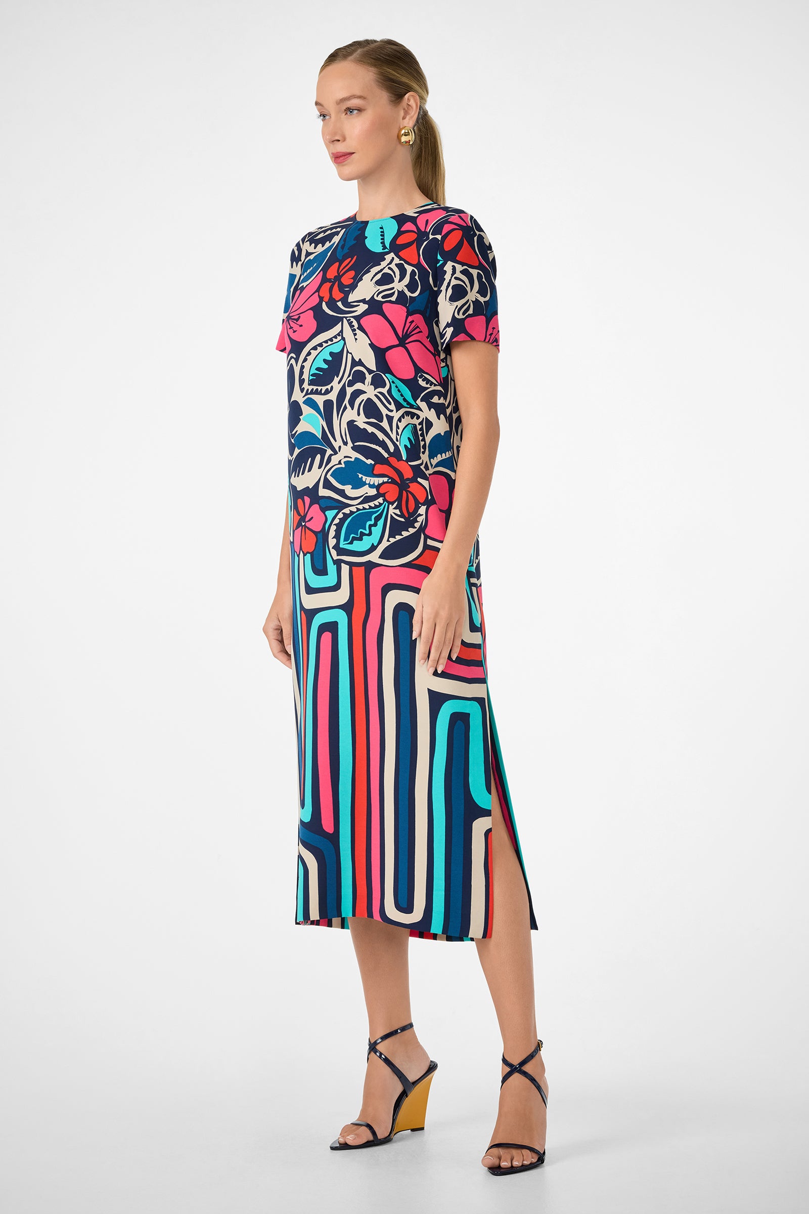 A woman wears the Martay Dress, a colorful short-sleeved piece with bold floral and geometric patterns in red, blue, and cream. She has blonde hair tied back and pairs it with strappy wedge sandals.