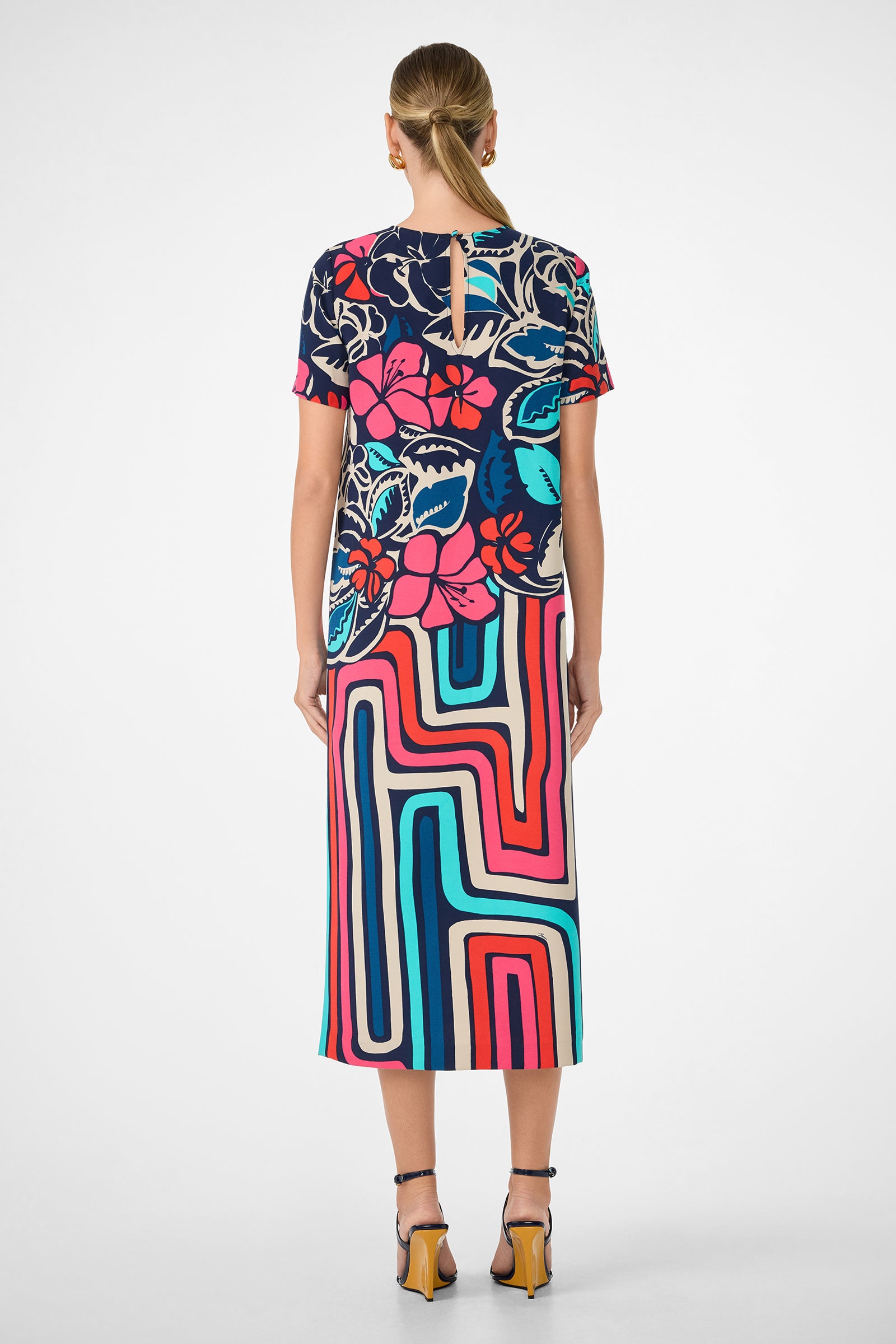 A woman with blonde hair in a low bun wears the Martay Dress, featuring a bold floral top and geometric striped skirt. She pairs it with strappy heels and stands against a plain white background, facing away from the camera.