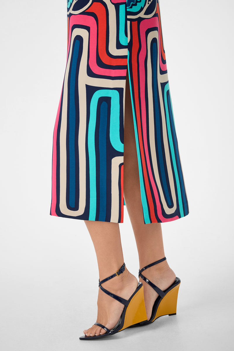 A person wearing the Martay Dress, which features a colorful, geometric pattern and side slit, paired with black strappy wedge sandals with yellow soles, stands against a light background.