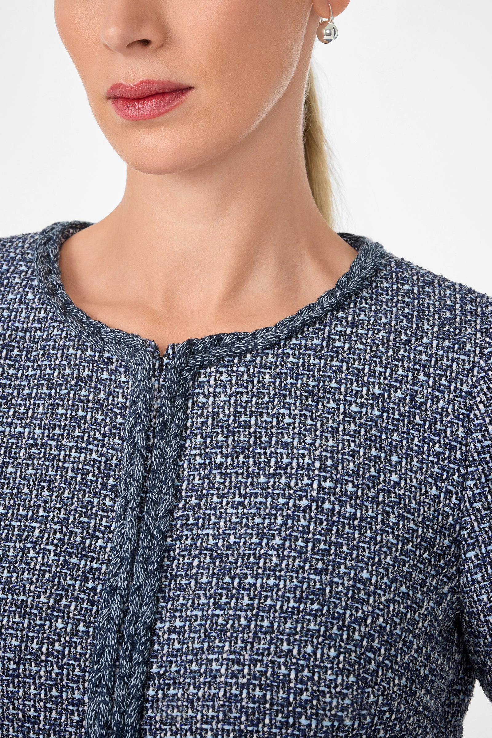 A woman with light skin wears the Kiui Blazer, a textured navy and white tweed jacket featuring a rounded neckline and subtle trim. She has pink lipstick, small hoop earrings, and only her face and upper torso are visible.