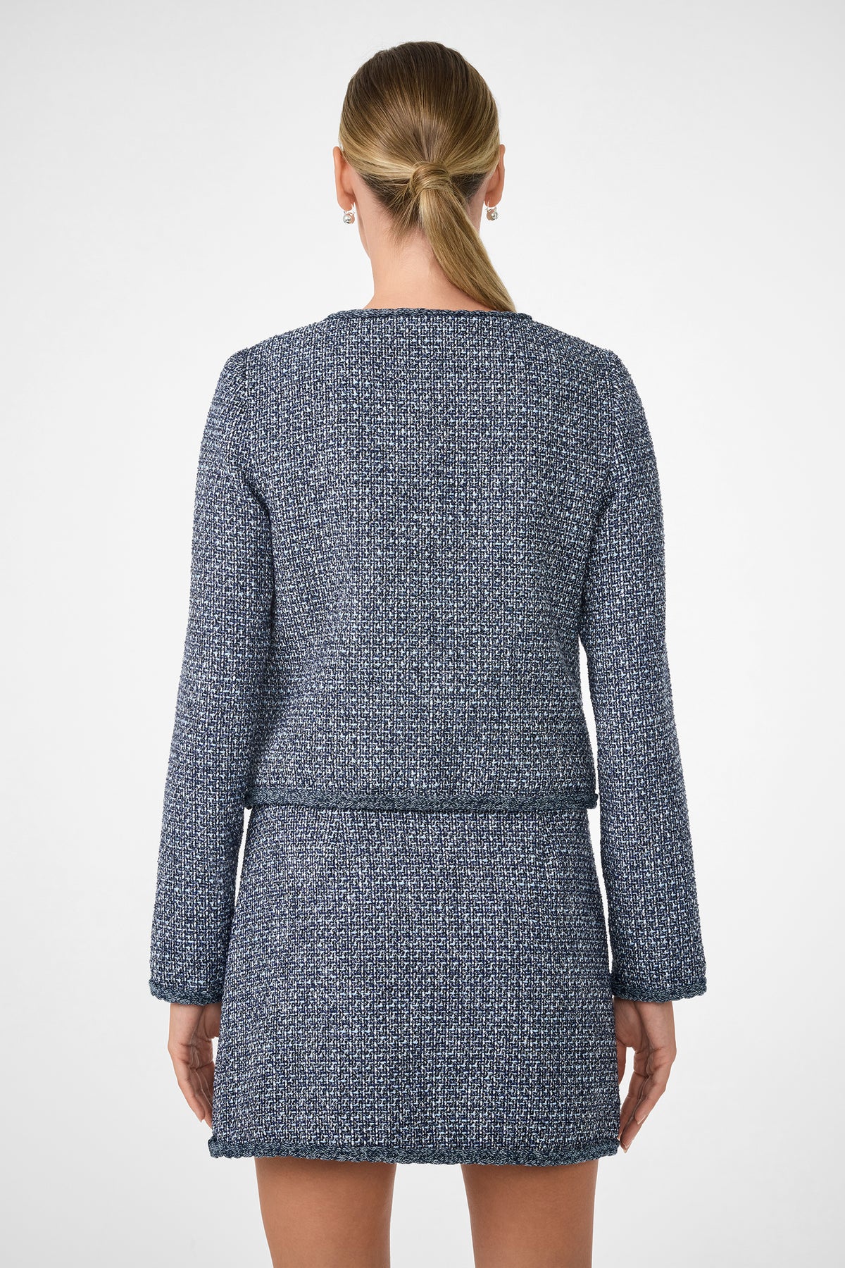 A woman with blonde hair in a low ponytail is shown from the back wearing the Kiui Blazer, a textured blue and white long-sleeve jacket with a matching short skirt, against a plain white background.
