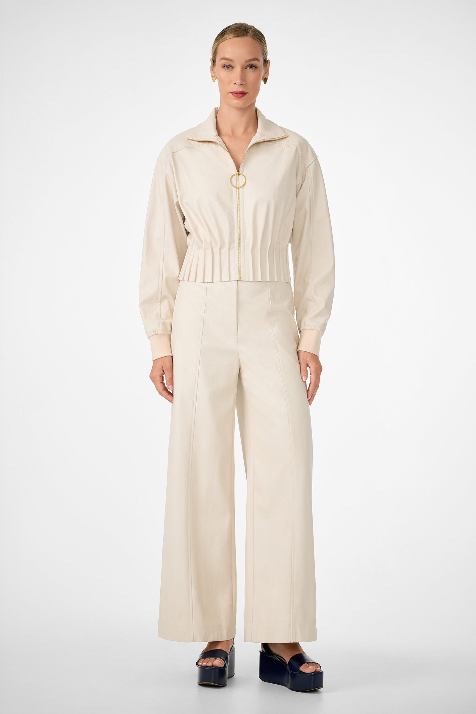 A woman stands against a white background wearing the stylish cream Outrun Jacket—a zip-up, long-sleeve top—paired with wide-leg pants. Her hair is slicked back and she completes the look with black platform sandals.