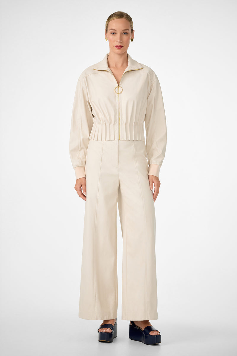 A woman stands against a white background wearing the stylish cream Outrun Jacket—a zip-up, long-sleeve top—paired with wide-leg pants. Her hair is slicked back and she completes the look with black platform sandals.