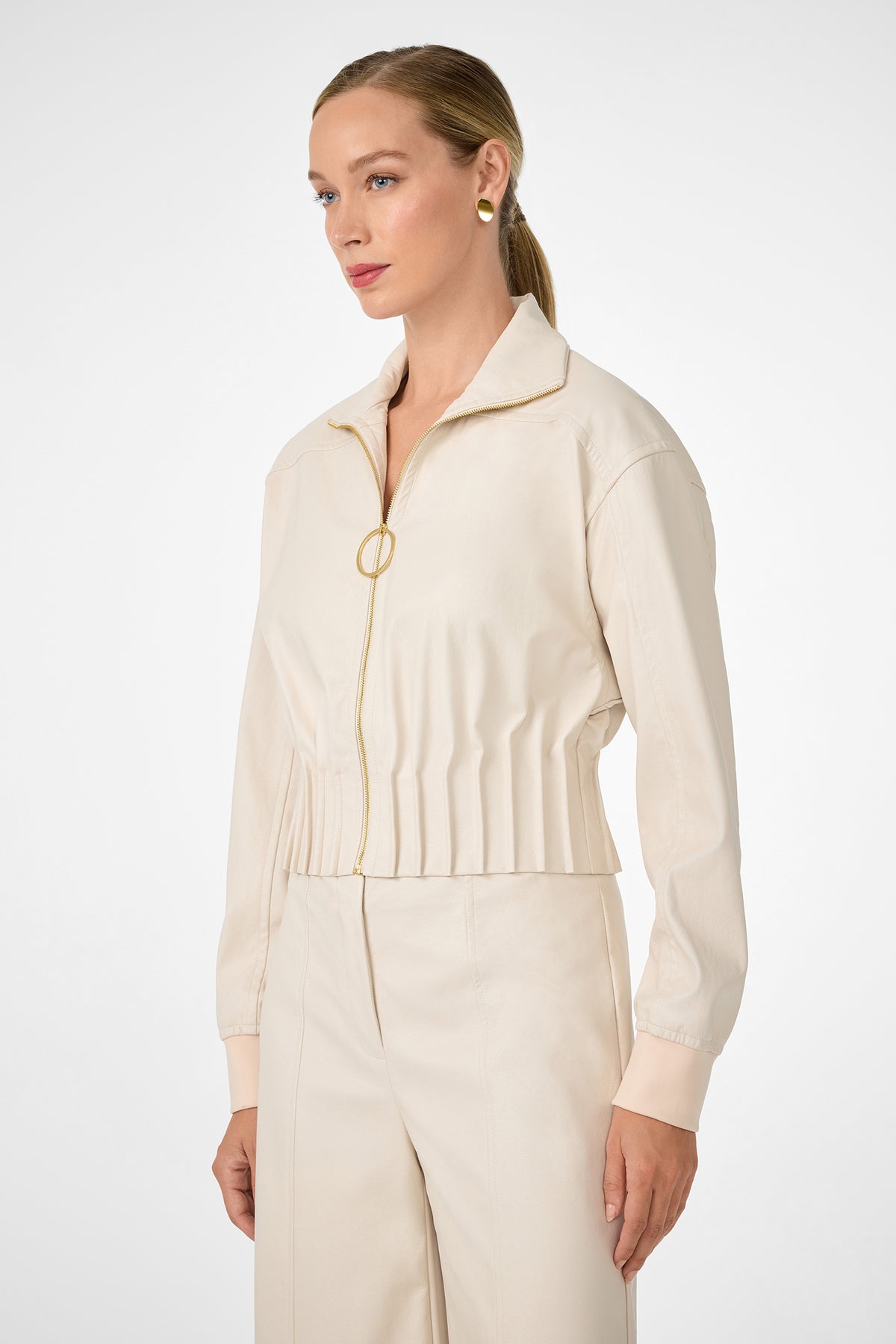 A woman with blonde hair in a low ponytail wears the cream Outrun Jacket with matching high-waisted pants, standing against a plain white background and looking to the side.