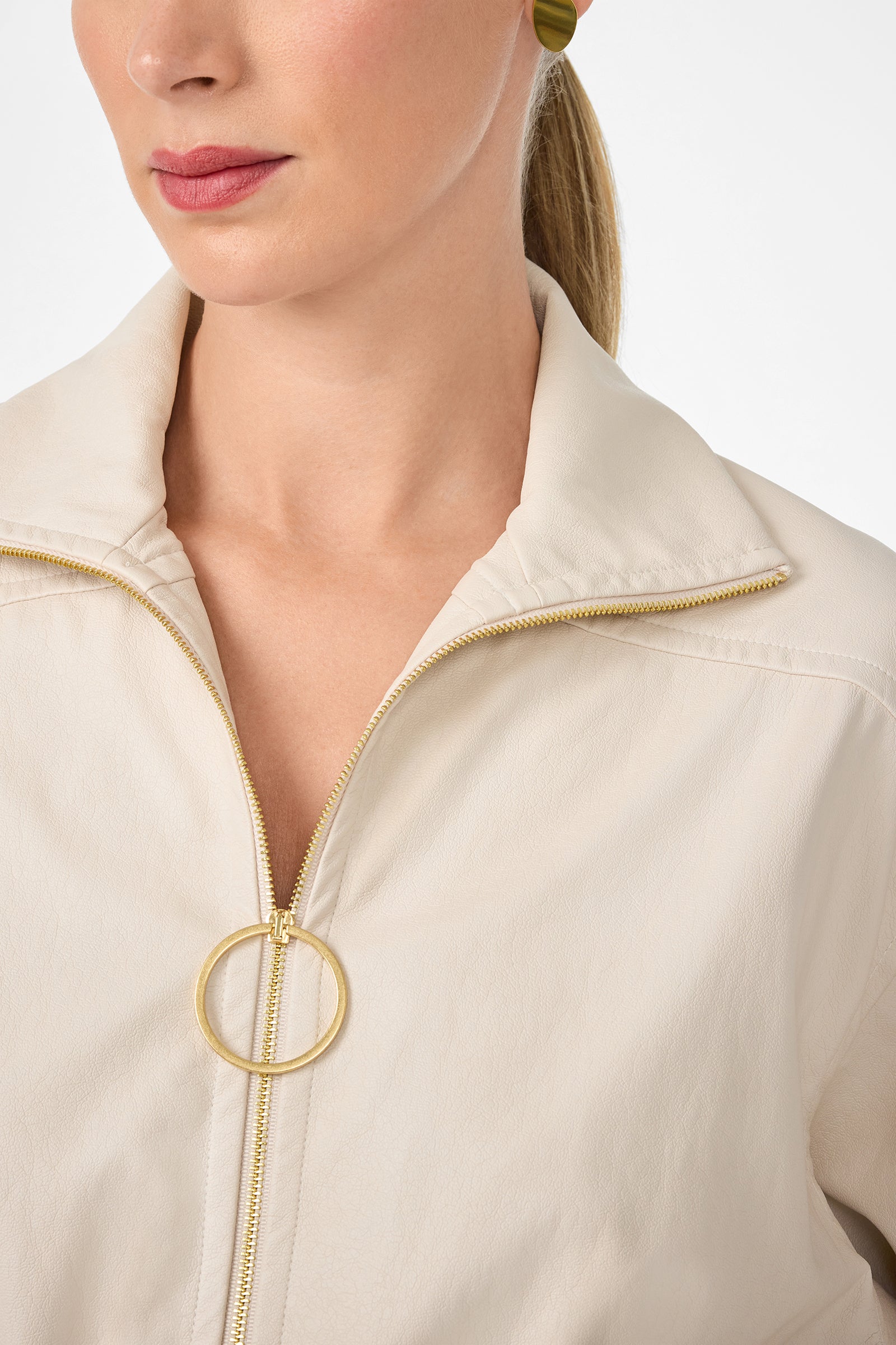 A woman with light skin and pink lipstick wears the cream Outrun Jacket featuring a gold circular zipper pull. Her blonde hair is tied back, she wears a green earring, and her face appears partially against a plain background.