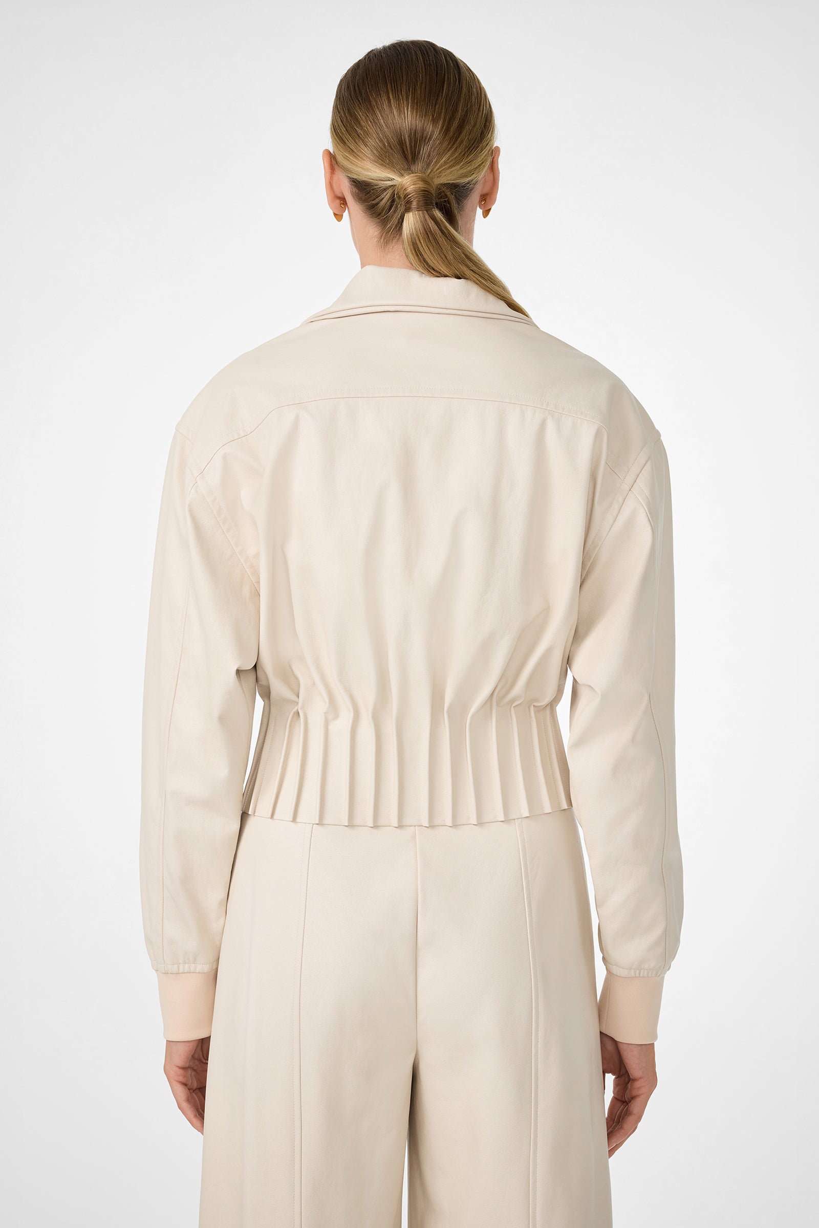 A woman with light brown hair in a low ponytail is seen from behind, wearing the cream-colored Outrun Jacket with a cinched waist and matching high-waisted pants against a plain light background.