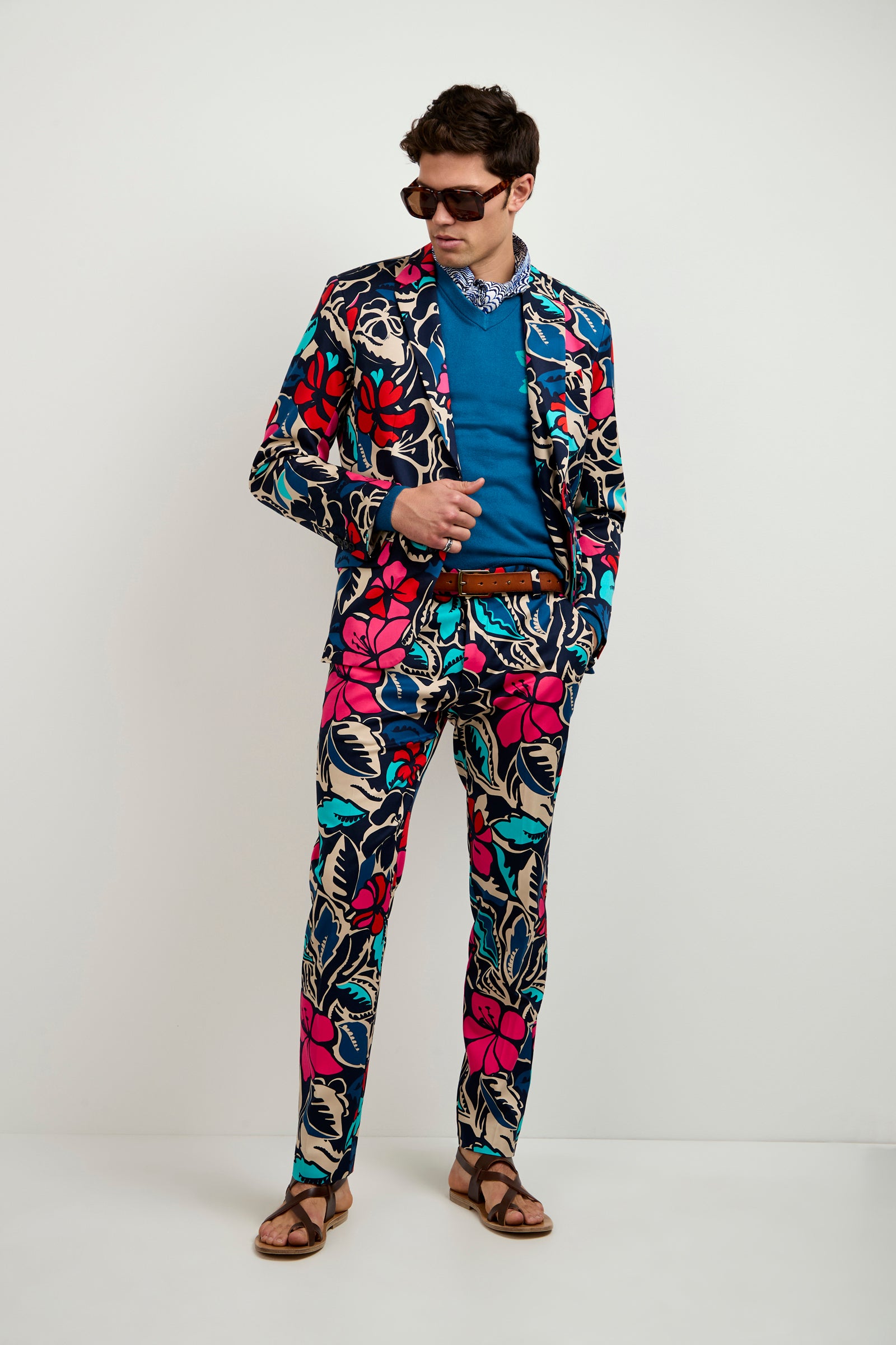 A man in sunglasses wears the Thurston Blazer - Copacabana, featuring a vibrant floral pattern in blue, pink, and cream over a blue shirt with a brown belt and sandals, all set against a plain white background.