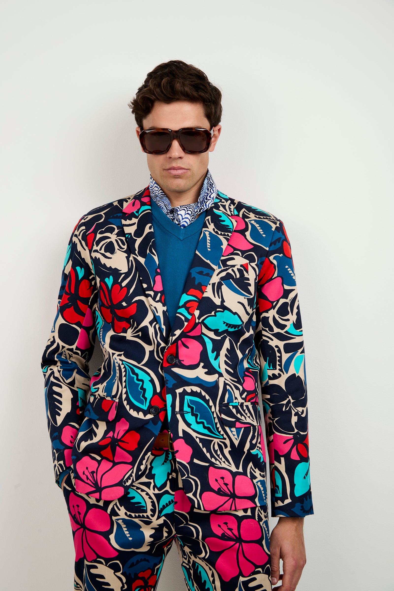 A man wearing dark sunglasses and the Thurston Blazer - Copacabana, featuring bold, colorful floral patterns, stands against a white background. He completes the look with a blue sweater and patterned scarf.