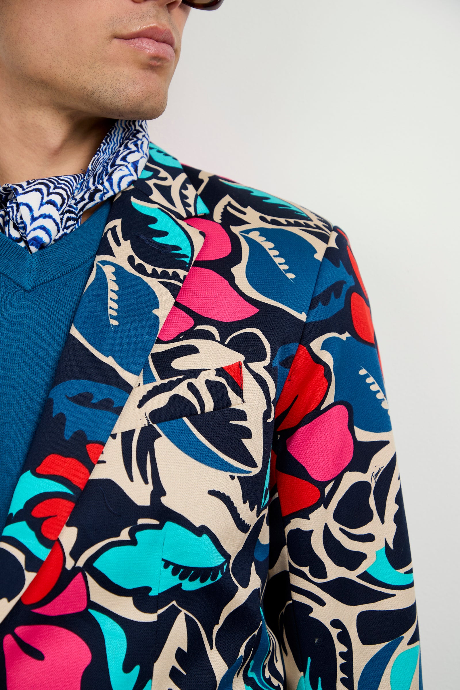 A person wearing the Thurston Blazer - Copacabana, featuring a bold floral and leaf print in red, teal, blue, and beige, layered over a blue sweater and a patterned blue and white shirt. Only the lower face and upper torso are visible.