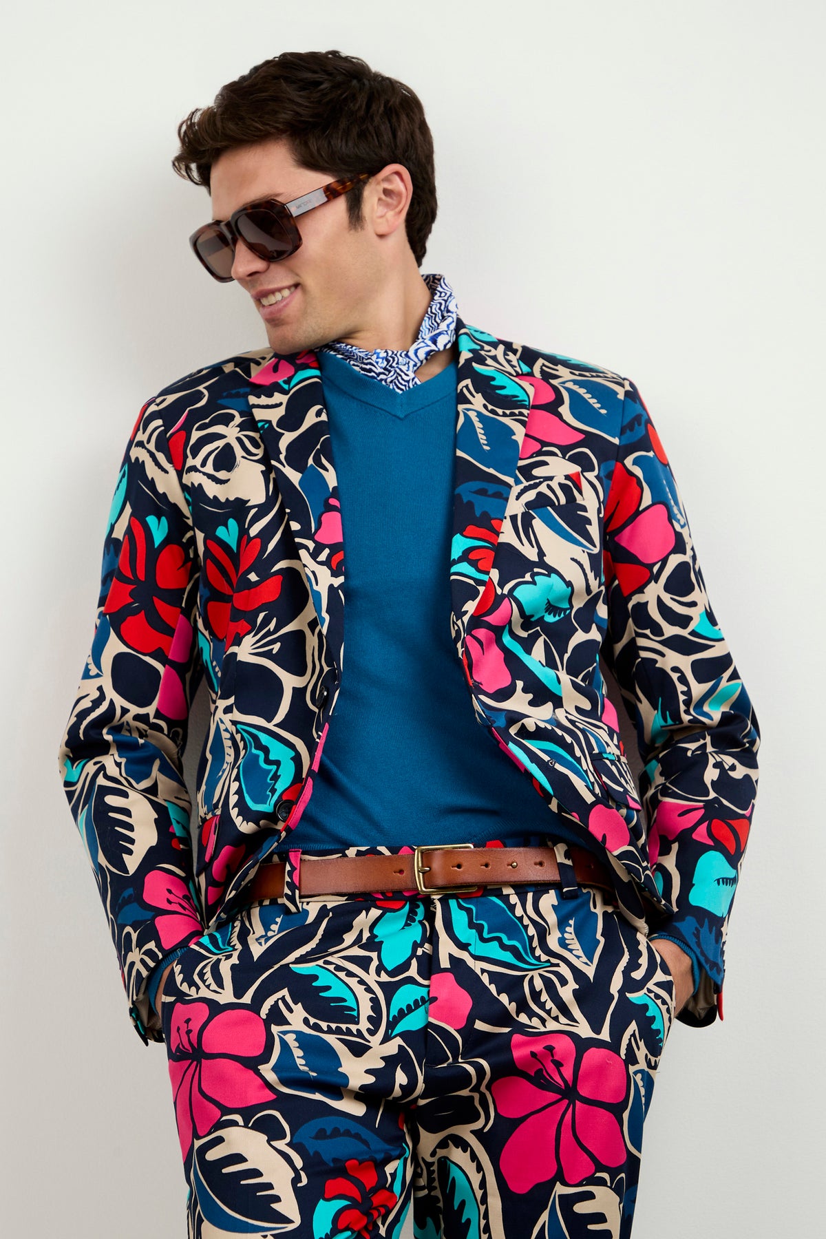 A man in large sunglasses wears the Thurston Blazer - Copacabana, featuring a bold tropical floral pattern in red, blue, teal, and tan over a blue sweater and collared shirt. He stands smiling against a plain white background.