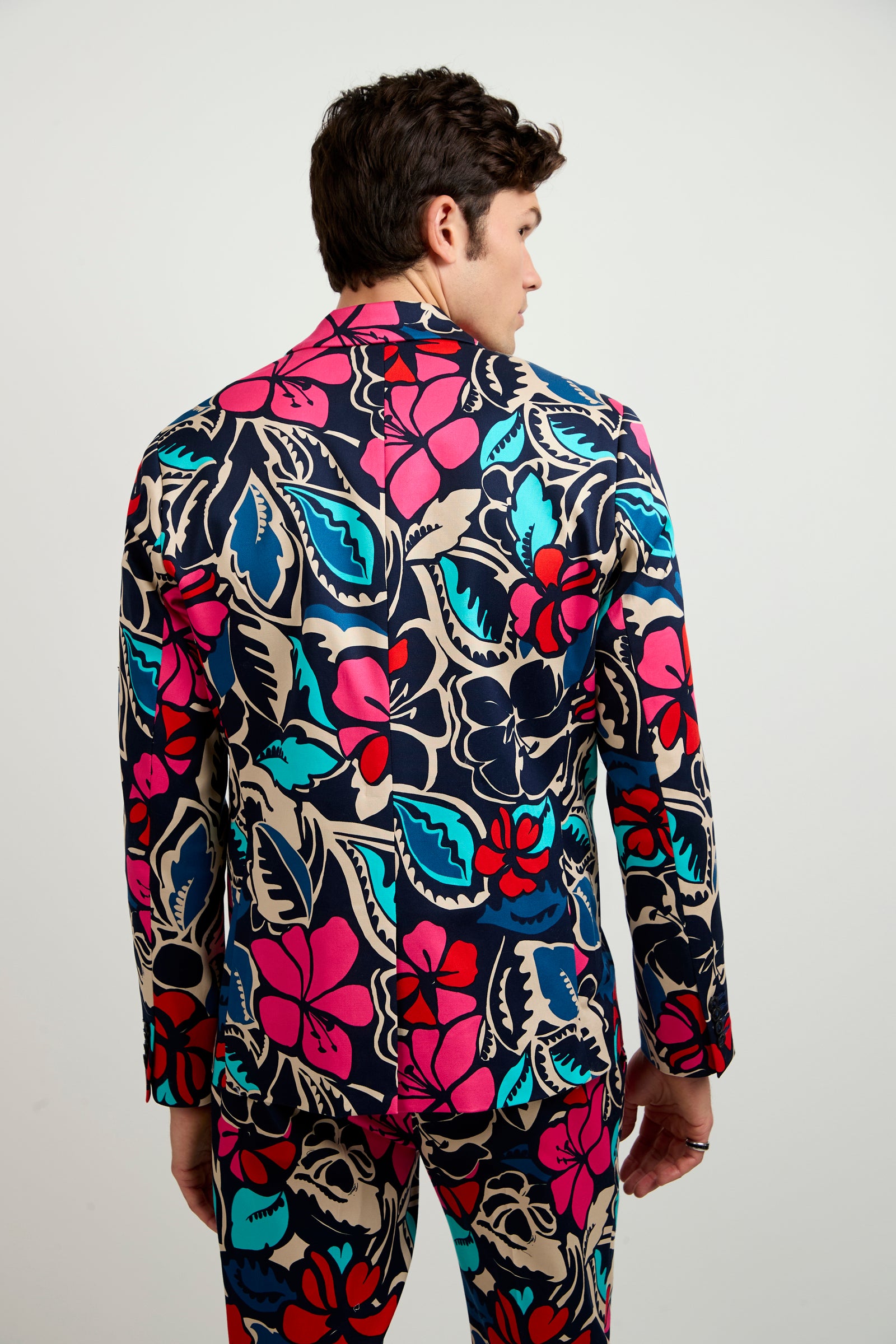 A person faces away, wearing the Thurston Blazer - Copacabana: a vibrant suit featuring a bold floral pattern in pink, red, teal, and blue hues on beige, paired with matching jacket and pants.