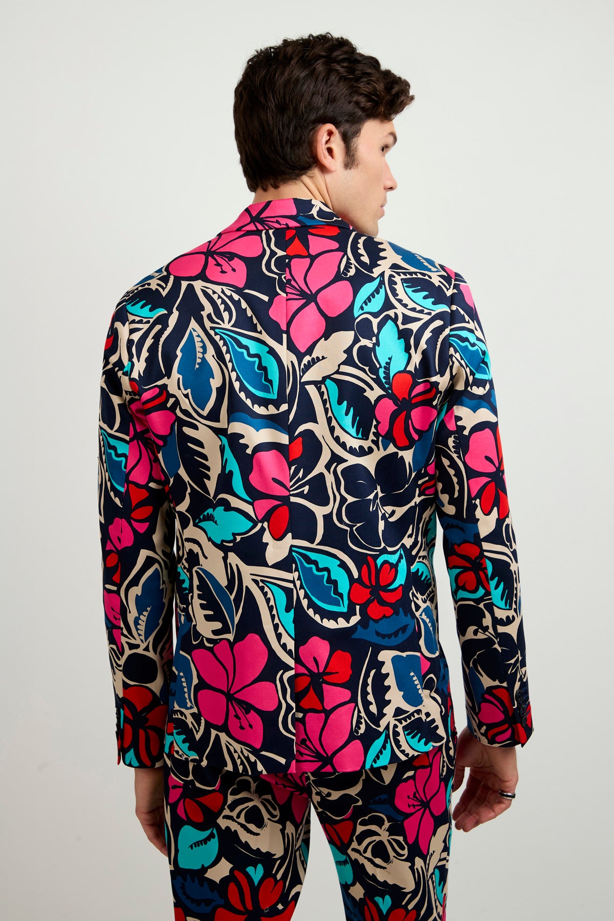 A person faces away, wearing the Thurston Blazer - Copacabana featuring a bold floral print in pink, blue, red, and beige on a light background. The man's hair is short and dark, set against a plain white backdrop.