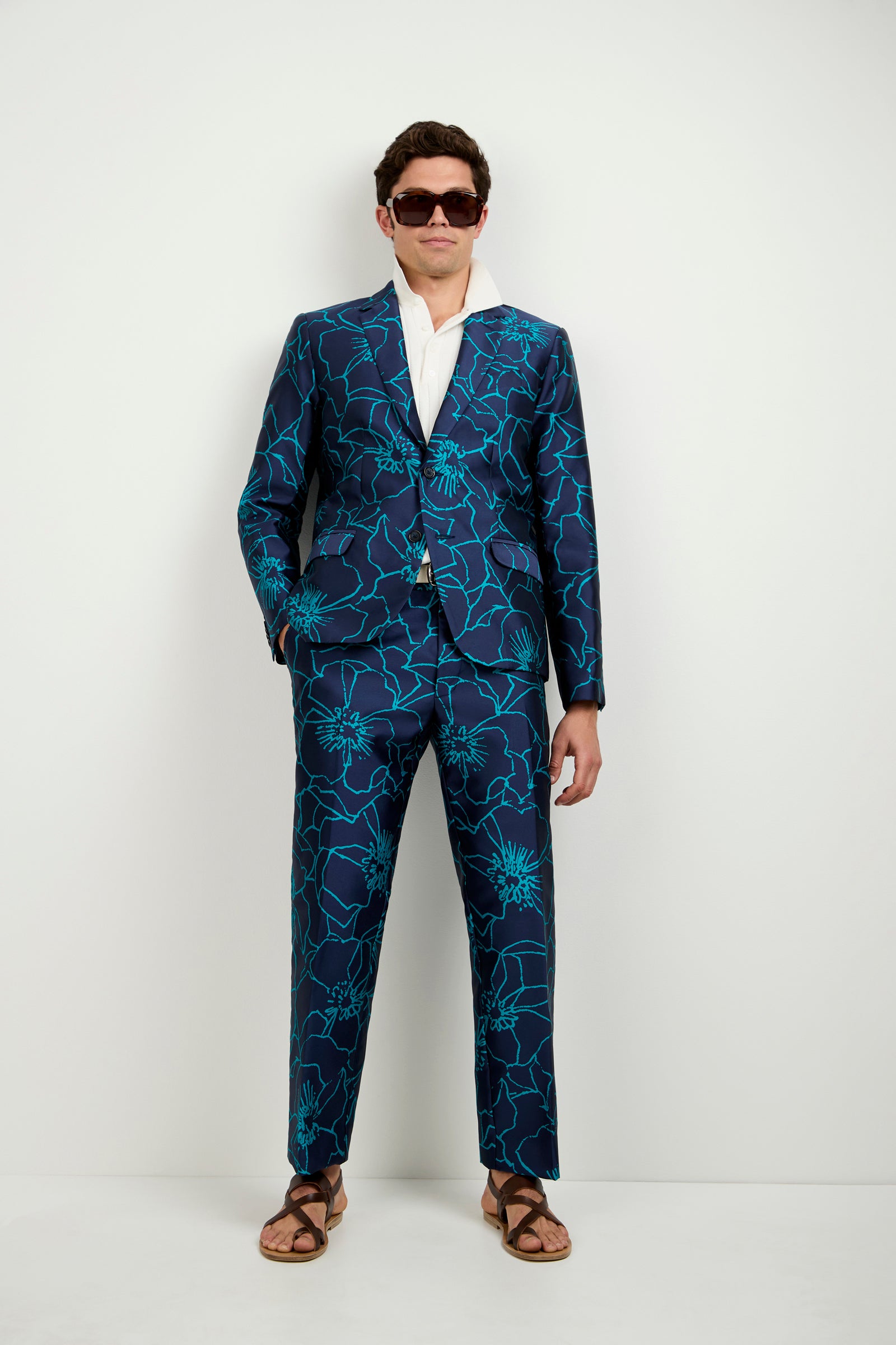 A man stands against a white wall wearing the Clyde Slim Trouser - Jucara Jacquard in a dark blue suit with turquoise floral pattern, paired with a white shirt, sunglasses, and brown sandals.
