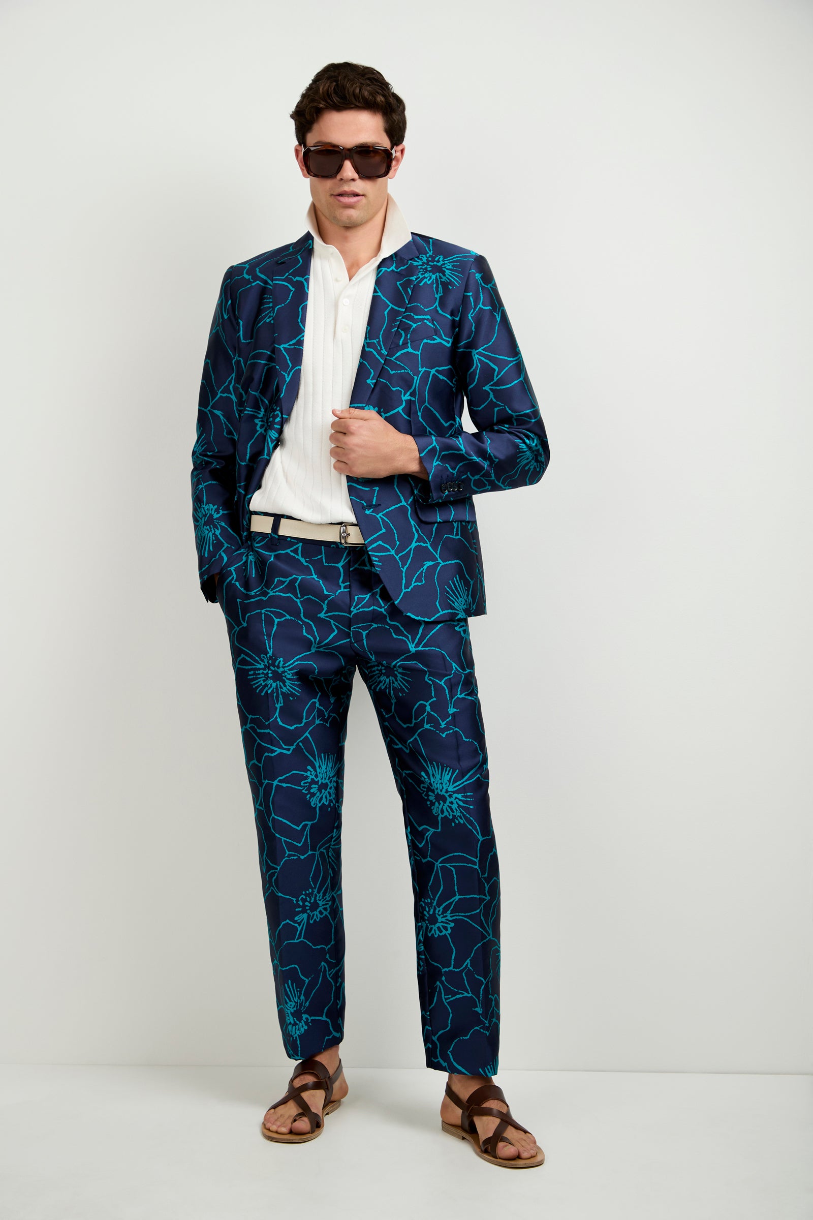 A man stands against a white background wearing sunglasses, a white shirt, brown sandals, and the Clyde Slim Trouser - Jucara Jacquard as part of his bold blue floral suit, with one hand resting on his jacket.