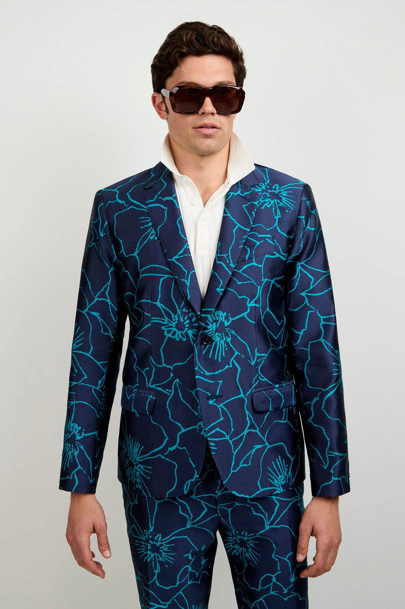Wearing the Thurston Blazer in Jucara Jacquard, featuring a bold turquoise floral pattern over blue, paired with a white shirt and large dark sunglasses, a person stands against a plain light background.