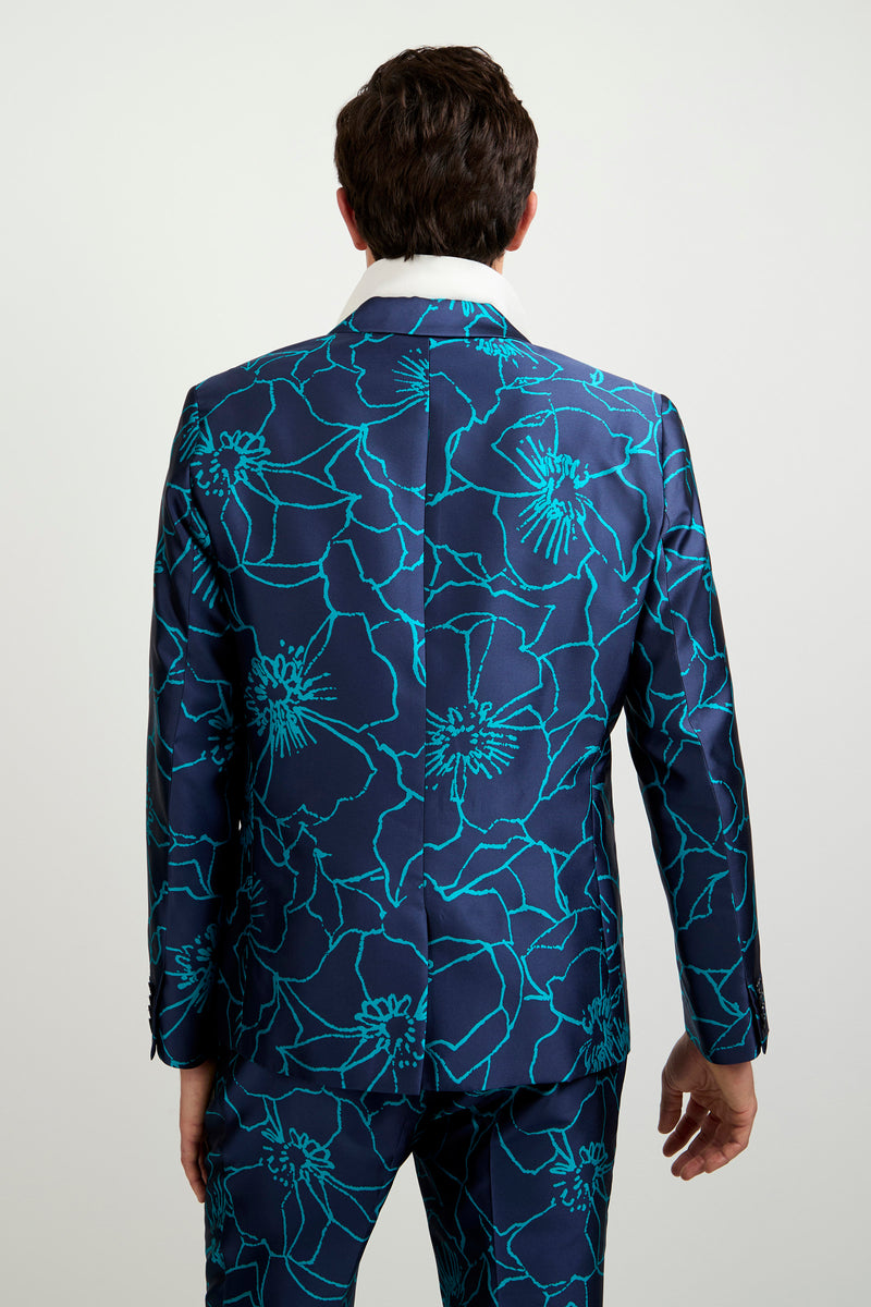 A person stands with their back turned, wearing the Thurston Blazer - Jucara Jacquard, featuring a shiny navy blue fabric with a striking turquoise floral pattern and a visible white shirt collar against a plain light background.