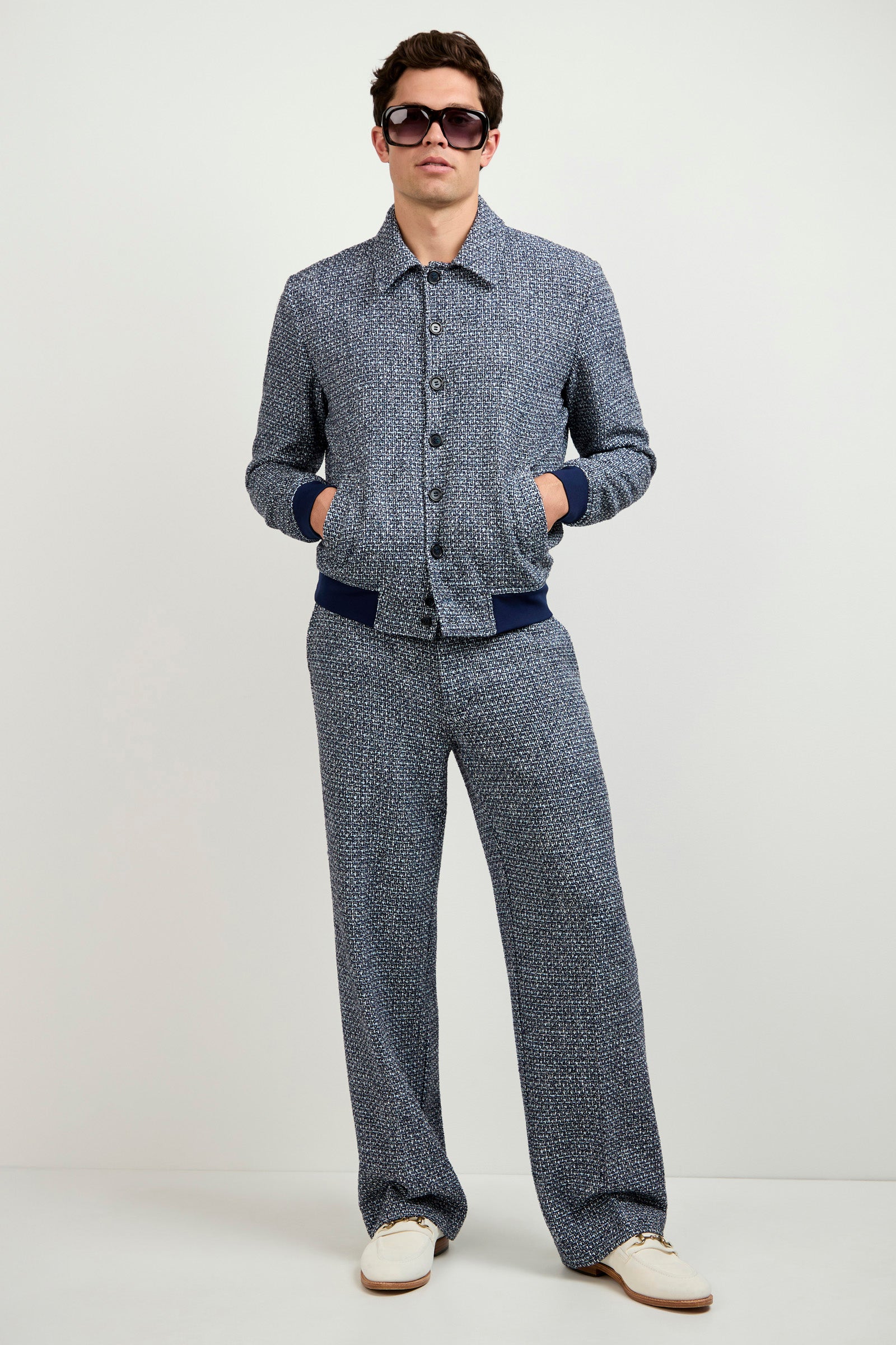 A person stands against a plain background, hands in pockets, wearing sunglasses, a blue patterned button-up jacket, Buckingham Tweed Trouser, and white shoes with beige soles.