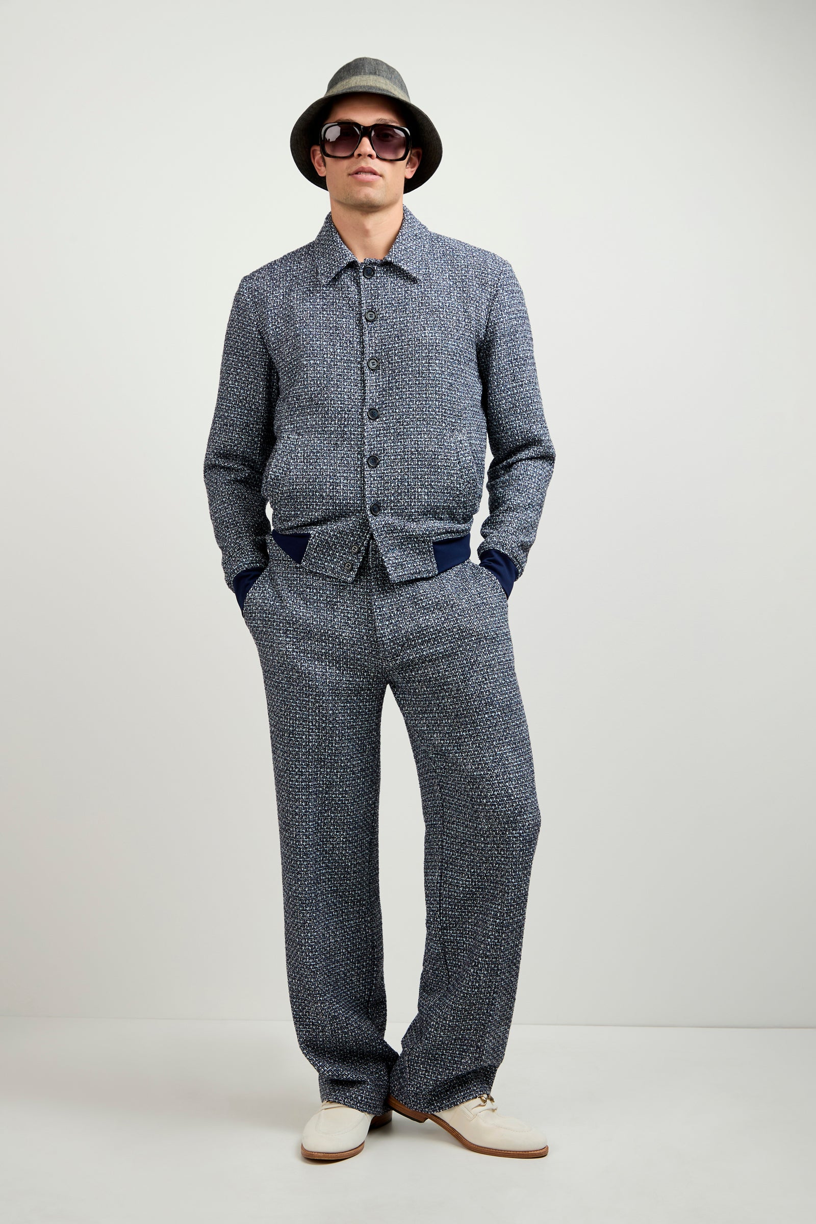 A man stands with his hands in his pockets against a plain light background, wearing the Lowen Tweed Jacket, matching pants, sunglasses, a gray hat, and white shoes.