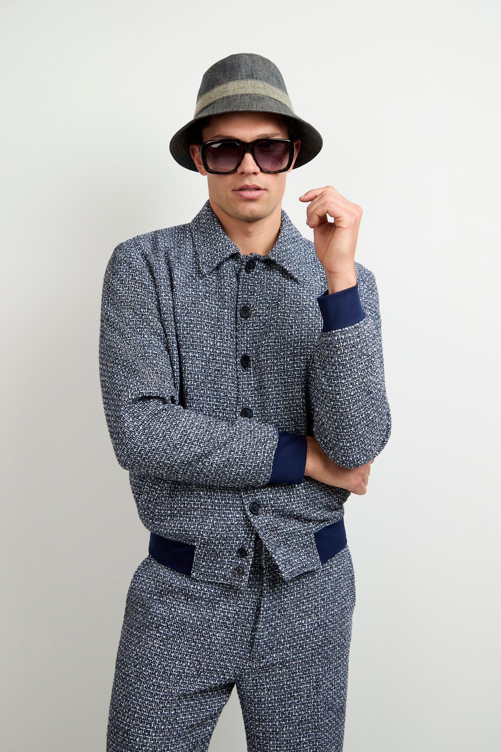 A person in a Lowen Tweed Jacket in Selva Suiting, paired with matching pants, dark sunglasses, and a gray fedora stands against a plain light background with one hand raised near their face.