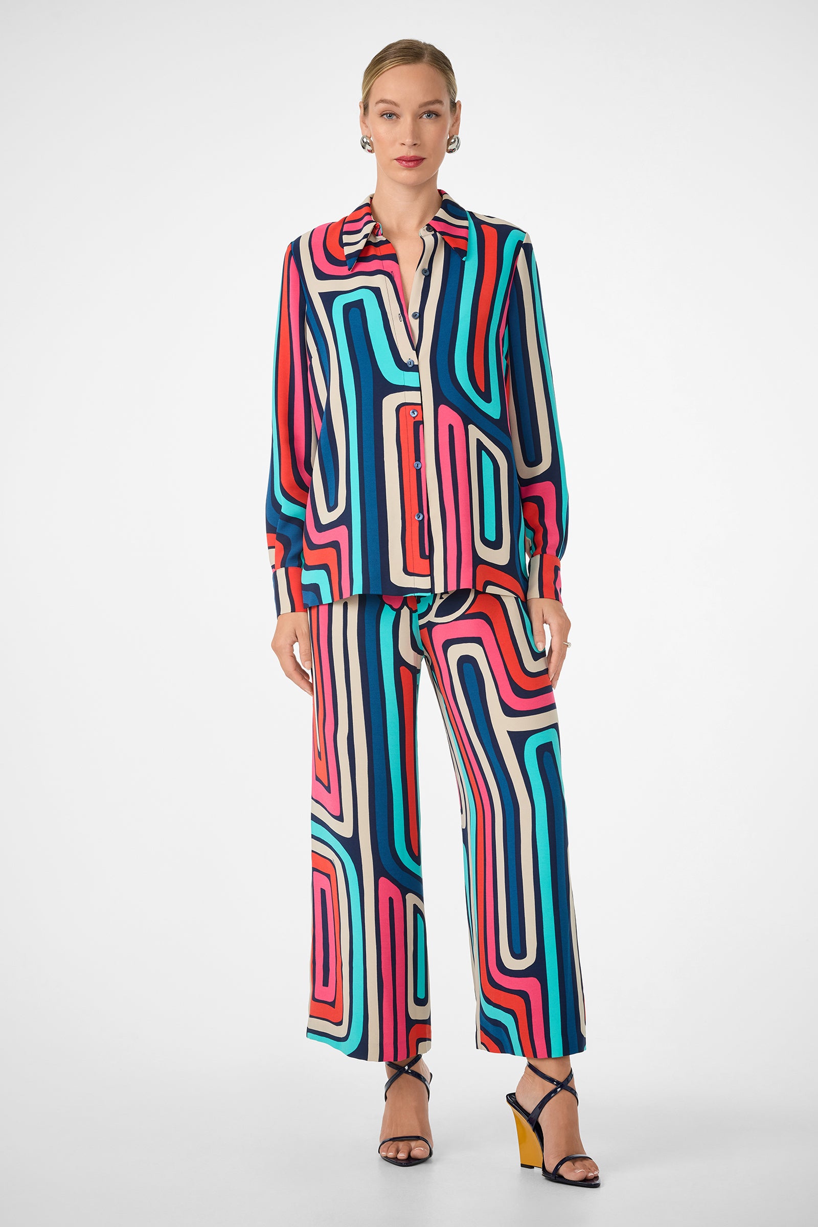 A person stands facing forward in the Cubism Pant, a colorful wide-leg matching set with a bold blue, pink, red, and black geometric pattern. The outfit includes a button-up shirt, paired with strappy heels and earrings.