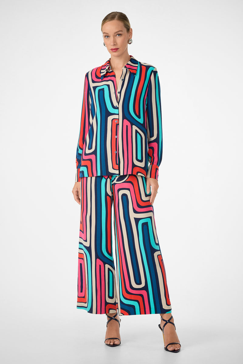 A woman wears the Montero Top, a long-sleeve button-up shirt featuring a bold geometric pattern in red, blue, pink, and black, paired with matching wide-leg pants and black strappy heels. Her hair is styled back.