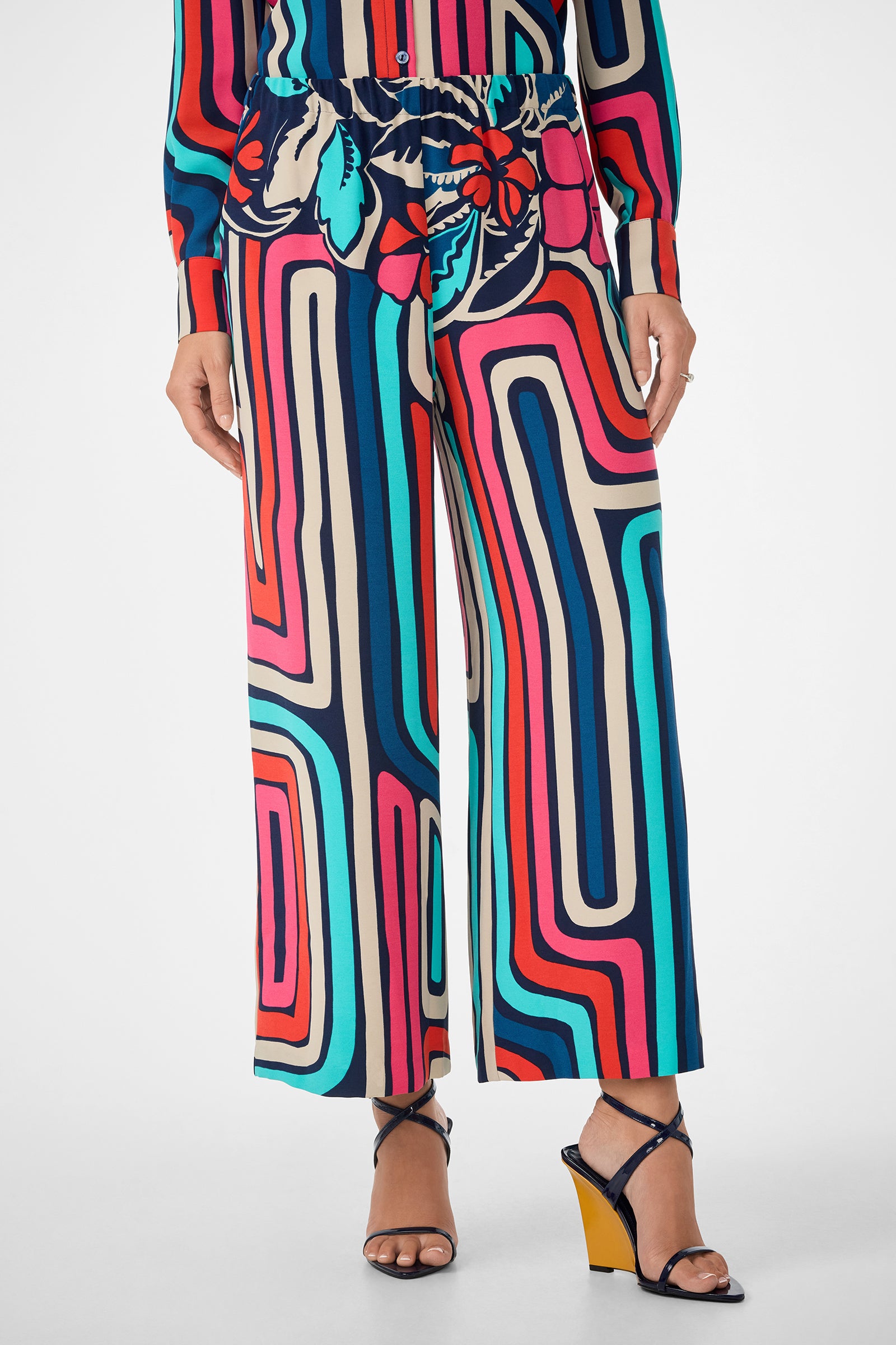 A person wears the Cubism Pant, featuring bold, colorful geometric and abstract prints in red, blue, teal, and pink tones, paired with a matching top and black strappy wedge sandals.