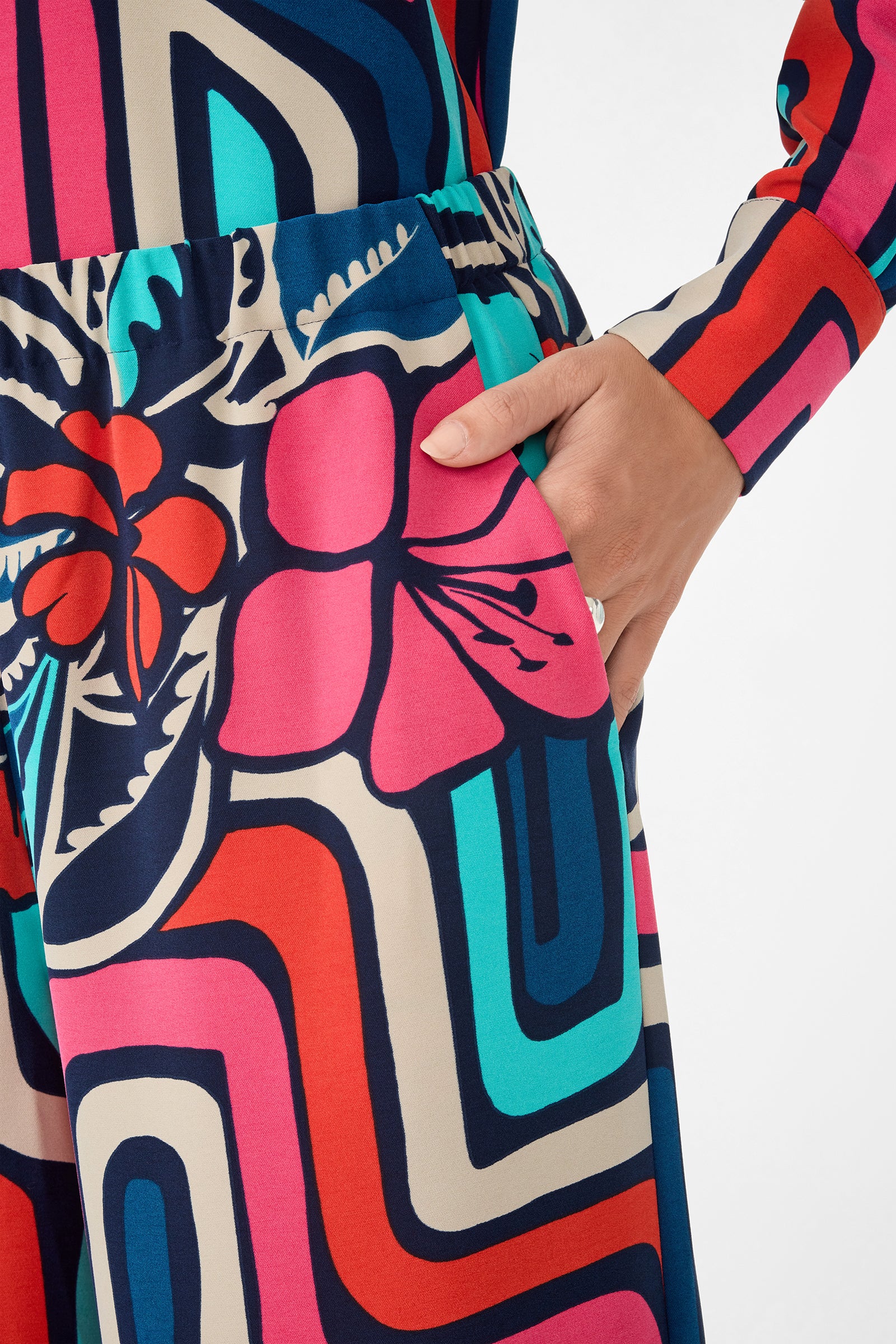 A hand is placed in the pocket of the Cubism Pant, which features bold floral and geometric patterns in pink, red, blue, teal, and cream on a plain white background.