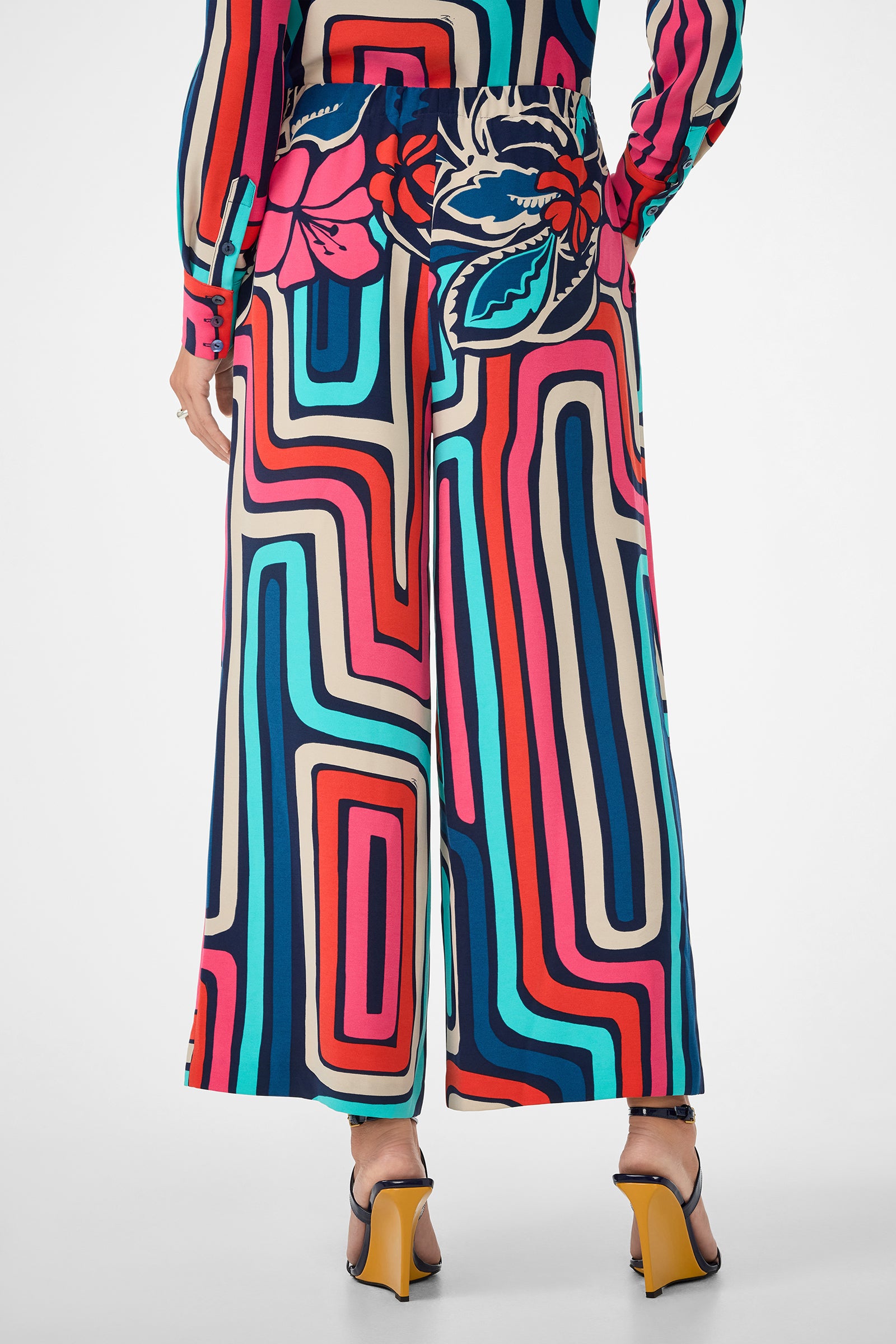 A person wearing the Cubism Pant, featuring a bold, colorful geometric and floral print in a wide-leg style, paired with high-heeled sandals. The image shows the back view from waist down.