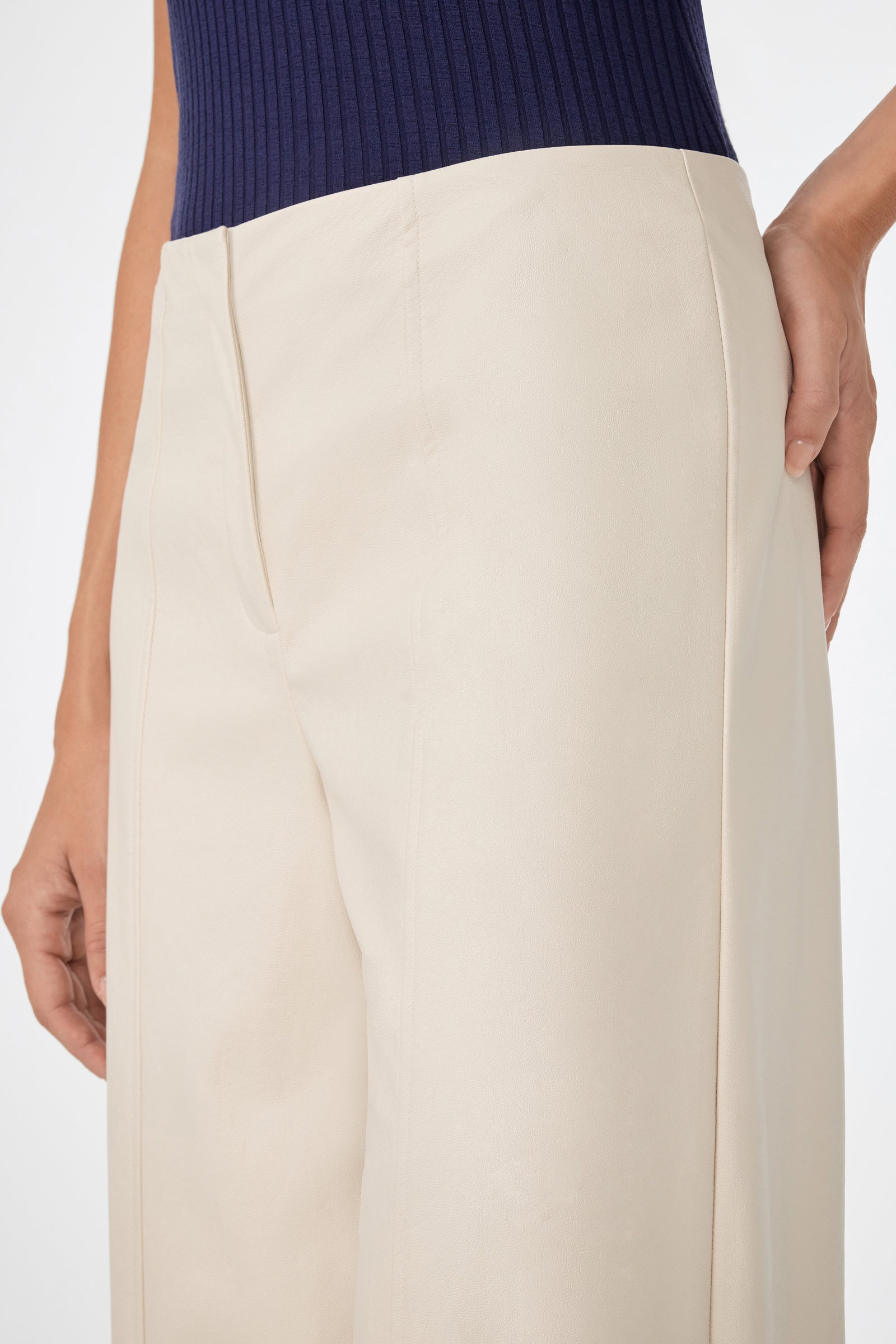 A person wearing the Parque Pant—high-waisted, wide-leg beige pants—paired with a sleeveless, ribbed navy top stands with one hand on their hip; the focus is on the pants and lower torso.