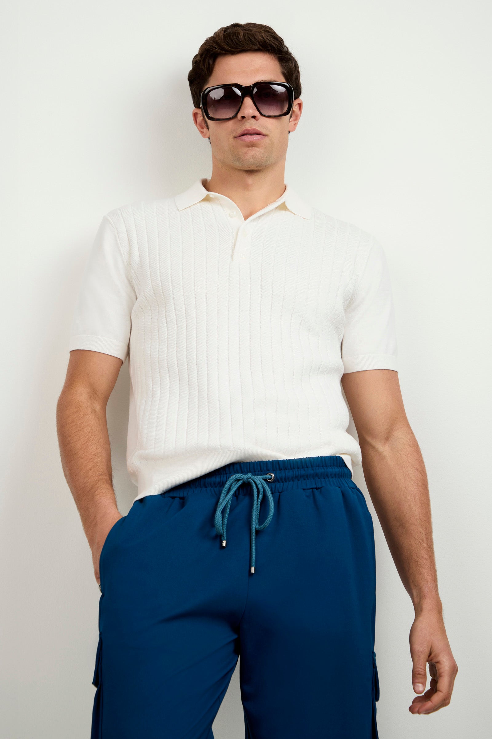 A man stands against a plain white background with one hand in his pocket, wearing dark sunglasses and the Alvarez Short-Sleeve Polo paired with blue drawstring pants.