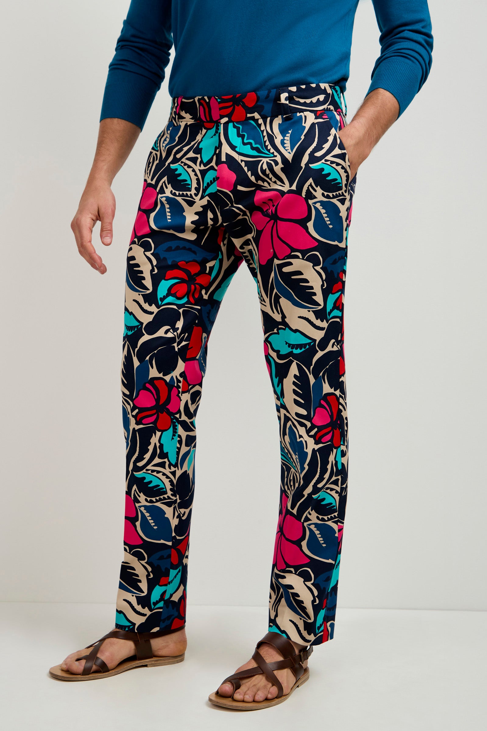 Someone wearing the Clyde Slim Trouser - Copacabana, featuring bold red, blue, teal, and beige floral prints, paired with a blue long-sleeve shirt and brown sandals, stands against a plain white background.