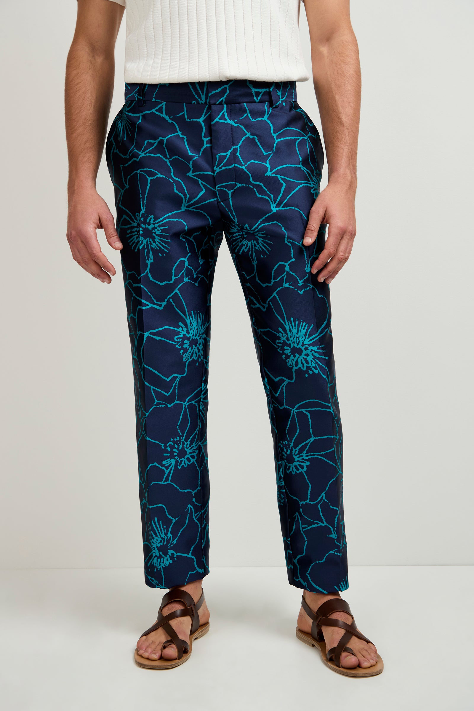 Wearing the Clyde Slim Trouser in Jucara Jacquard, a white ribbed short-sleeve shirt, and brown sandals, the person stands against a plain light background.