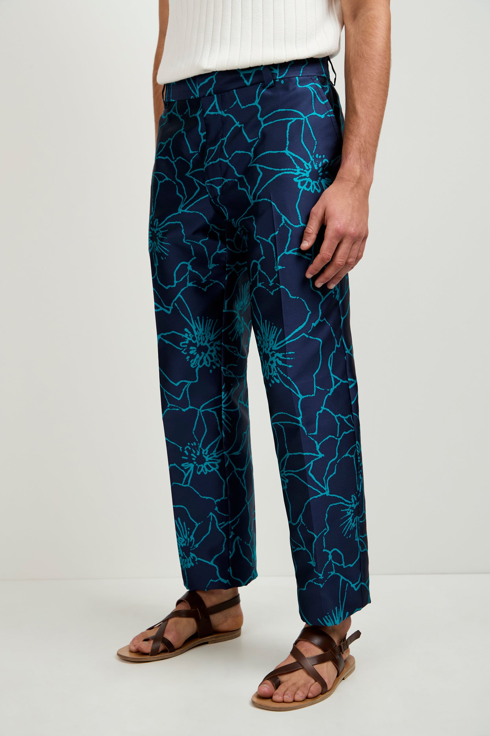 Wearing the Clyde Slim Trouser in Jucara Jacquard—a navy blue pant with a bold turquoise floral pattern—paired with a white ribbed top and brown strappy sandals, standing against a plain light background.