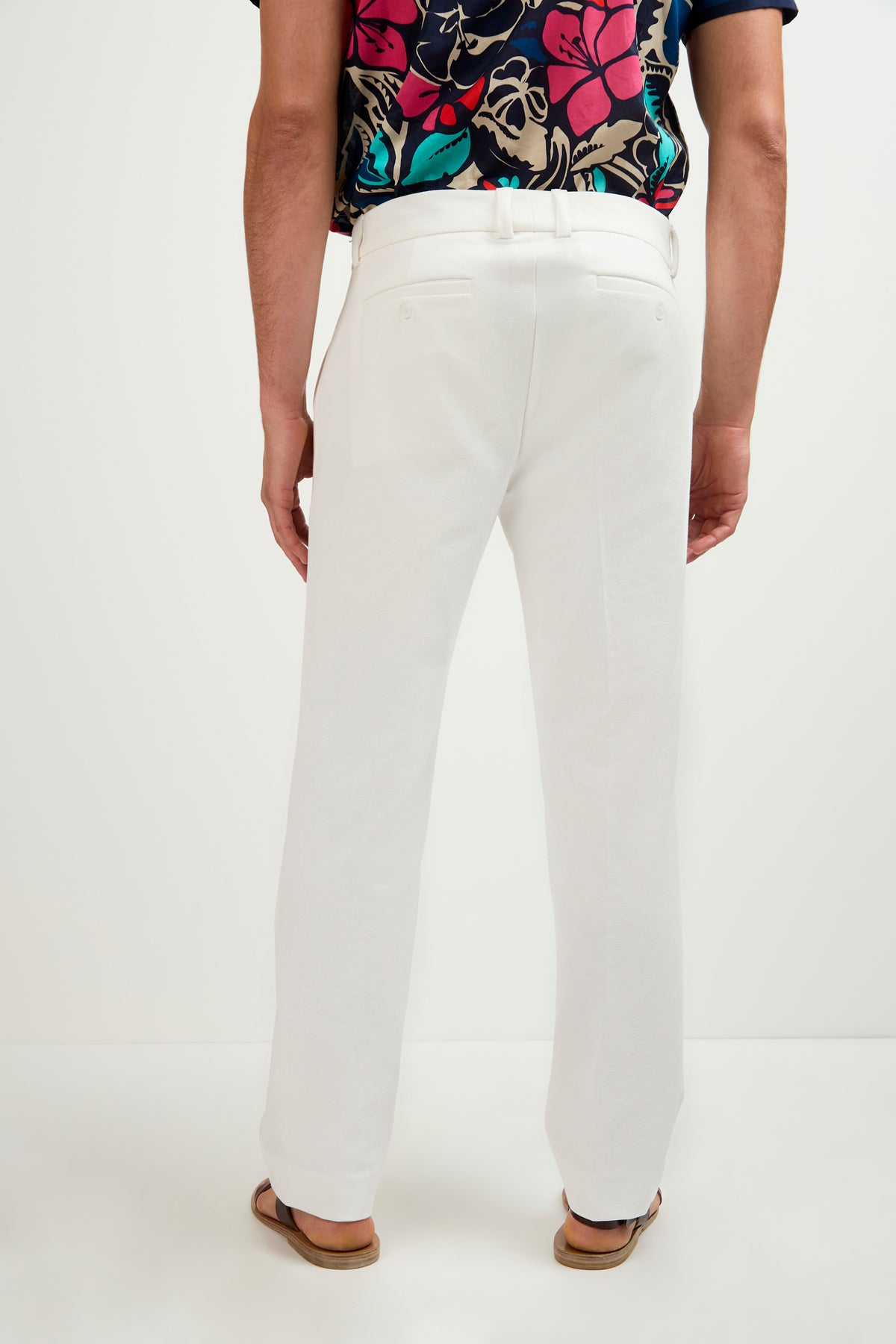 A person stands facing away, wearing Clyde Slim Trouser - Stretch Piqué in white, paired with a colorful floral shirt and sandals against a plain white background.